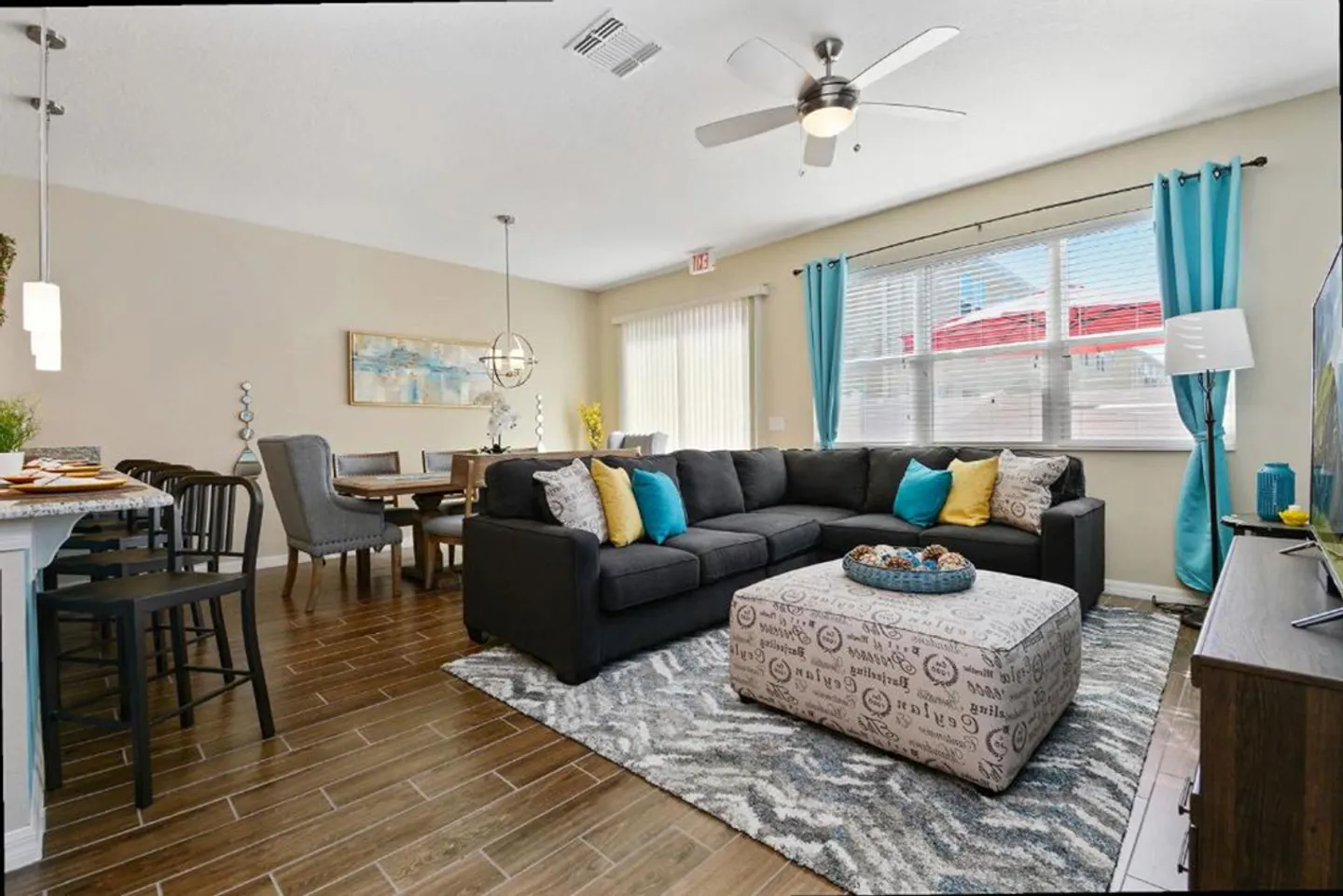 Open-concept living room featuring a dark gray sectional sofa, patterned ottoman, wood-look tile flooring, and adjacent dining area.