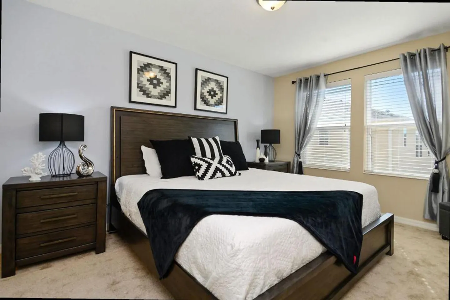 Bedroom featuring a dark wood bed with black and white bedding, flanked by nightstands and next to two windows with blinds.
