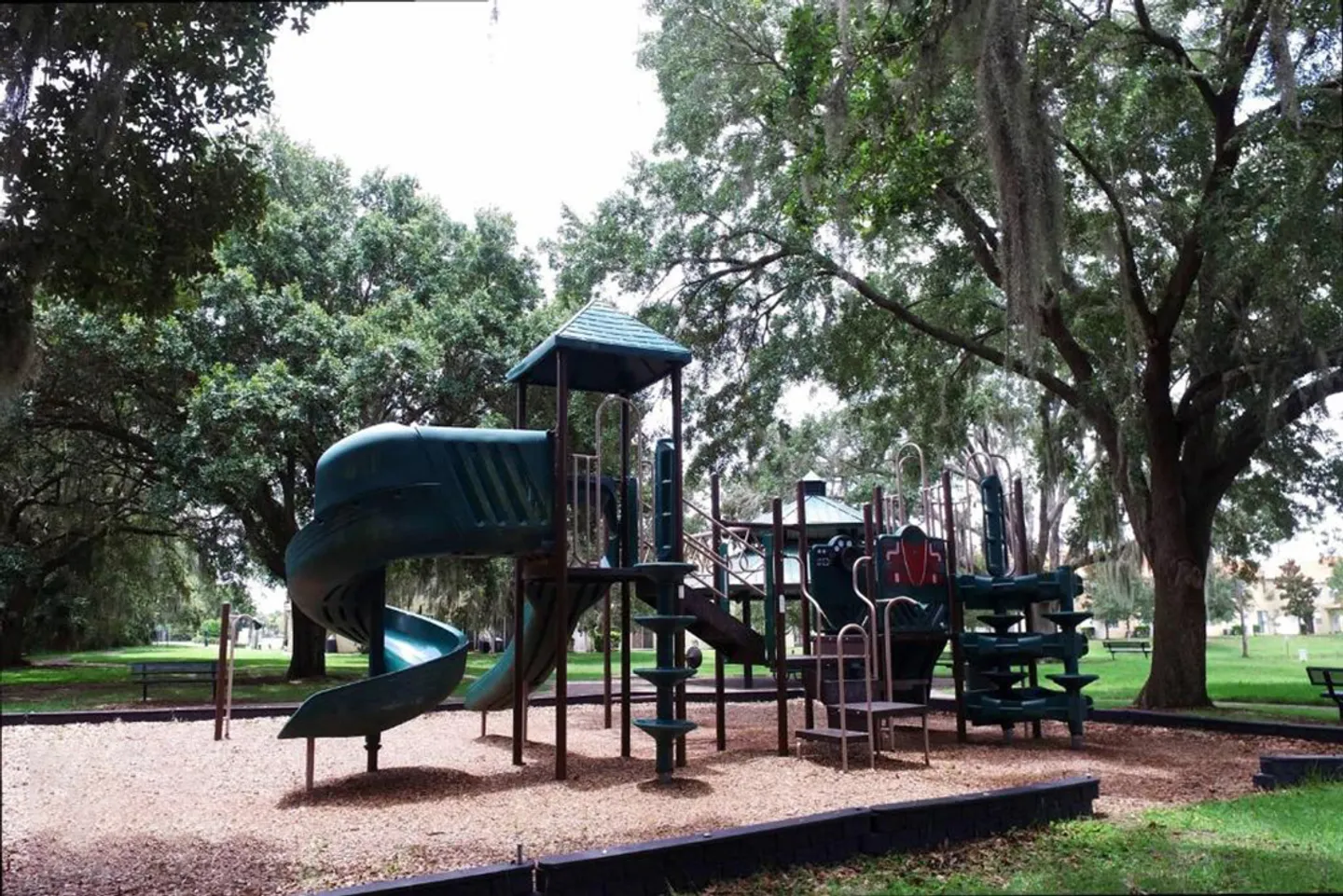 Outdoor children's playground structure with a green curved slide, platforms, and climbing steps surrounded by trees draped in Spanish moss.