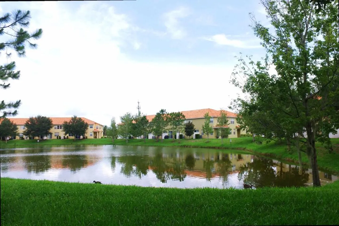 A scenic view across a grassy lawn and pond toward multi-story residential buildings with red tile roofs.