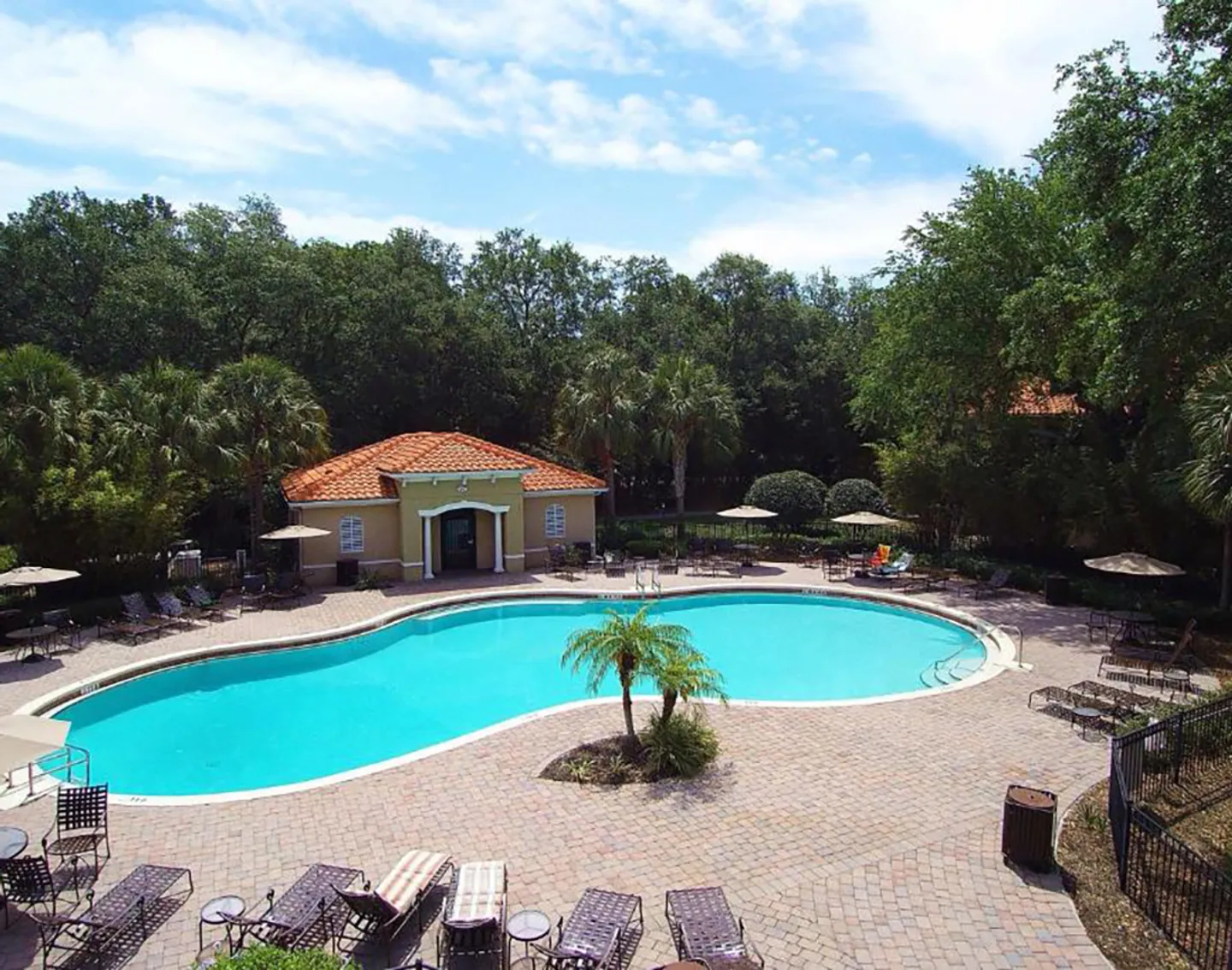 Elevated view of a curved outdoor community swimming pool surrounded by a brick paver deck, lounge chairs, and dense green trees under a partly cloudy sky.