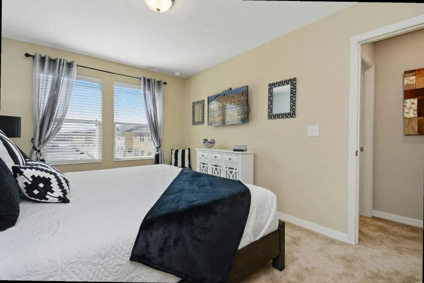 Bedroom with a bed in the foreground, two windows with blinds, a wall-mounted TV, and carpeted flooring.