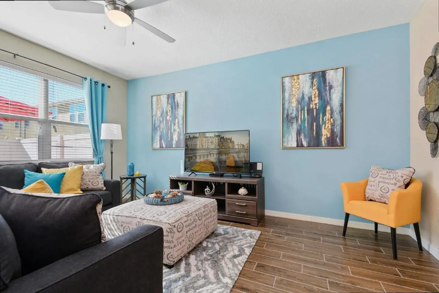 Living room with light blue accent wall, dark sectional sofa, printed ottoman, television on an entertainment console, and yellow armchair.