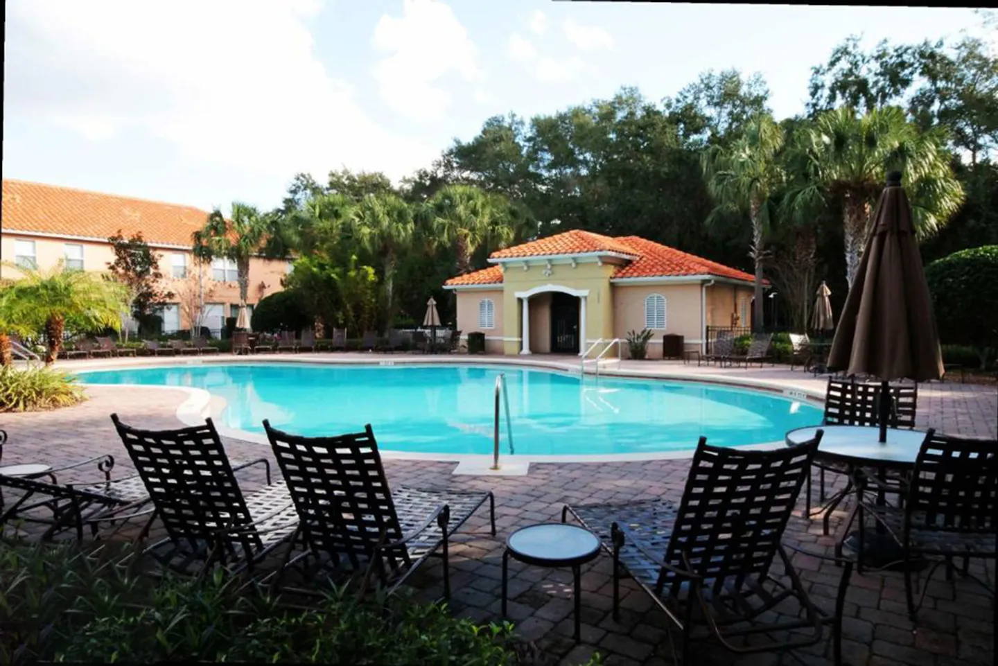 Community swimming pool surrounded by a brick paver patio, lounge chairs, palm trees, and a clubhouse with a red tile roof.