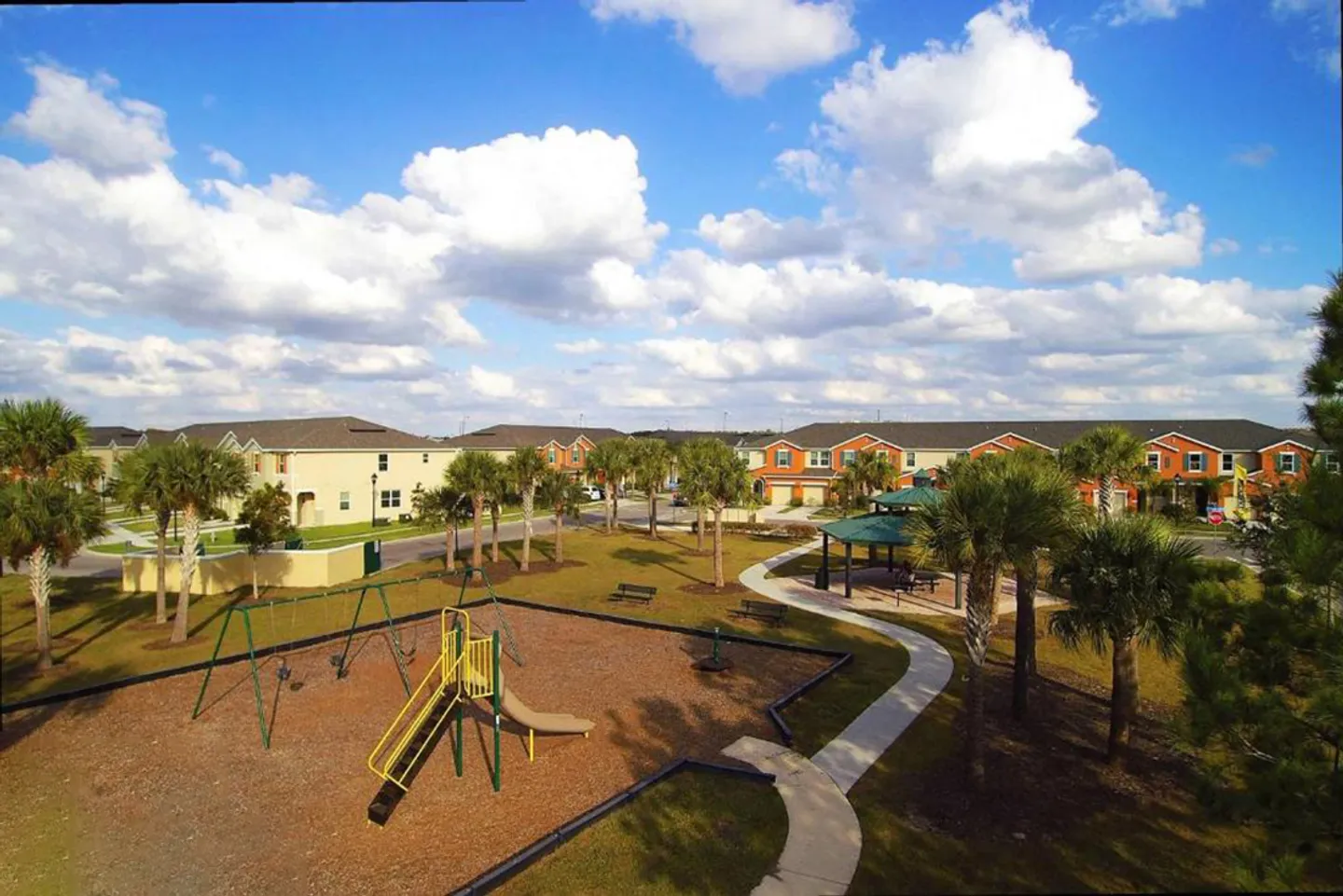 Elevated view of a community park with a playground, paved walking paths, covered gazebos, palm trees, and surrounding homes under a partly cloudy sky.