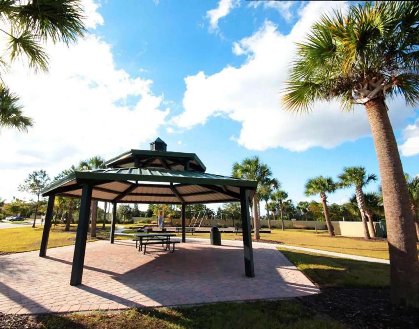A covered outdoor gazebo with picnic tables set on a brick patio in a community park with palm trees and a clear blue sky.
