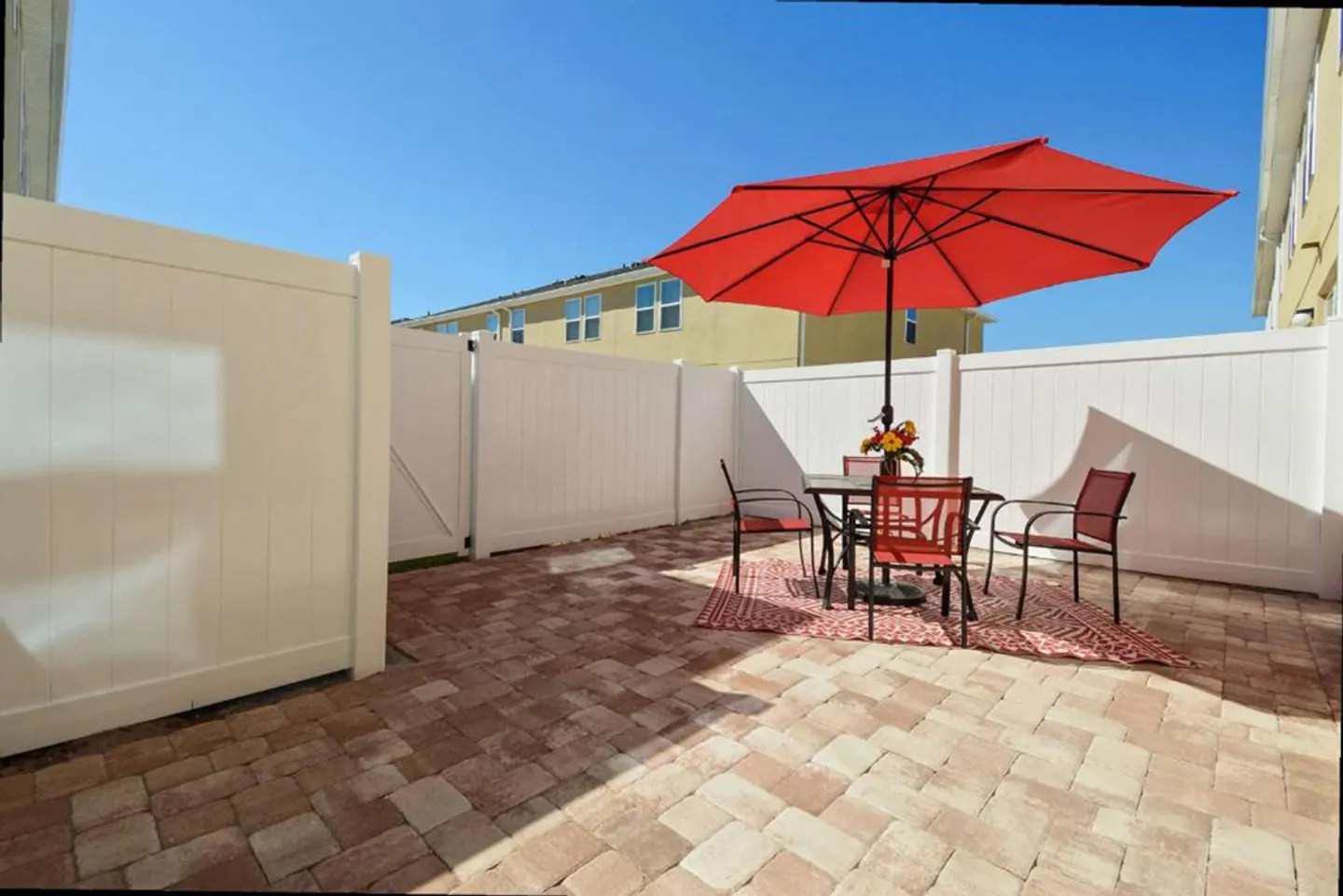 A paved outdoor patio enclosed by white vinyl fencing, featuring a patio table set with a red umbrella under a blue sky.