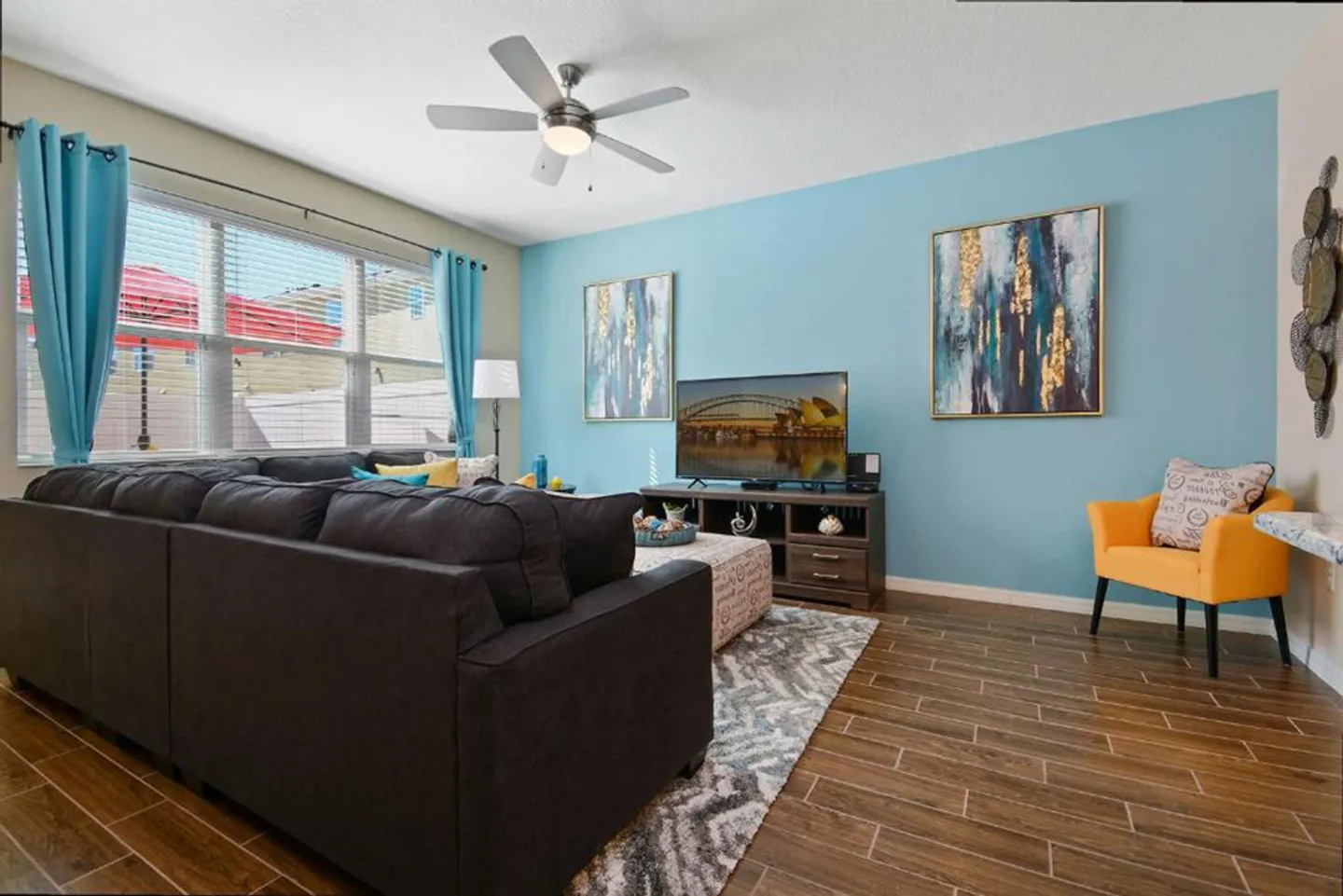 Living room with a large dark sectional sofa, wood-look tiled floors, a light blue accent wall with framed art, and a flat-screen TV.