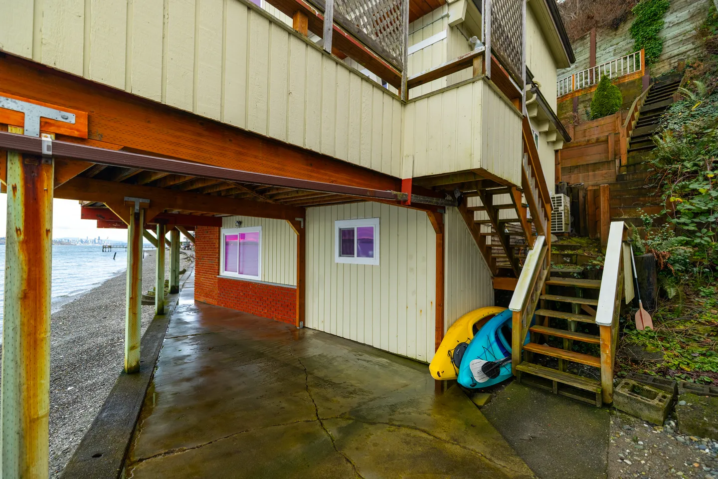 Exterior ground level under a raised waterfront home with concrete pad, kayaks under stairs, and shoreline access.