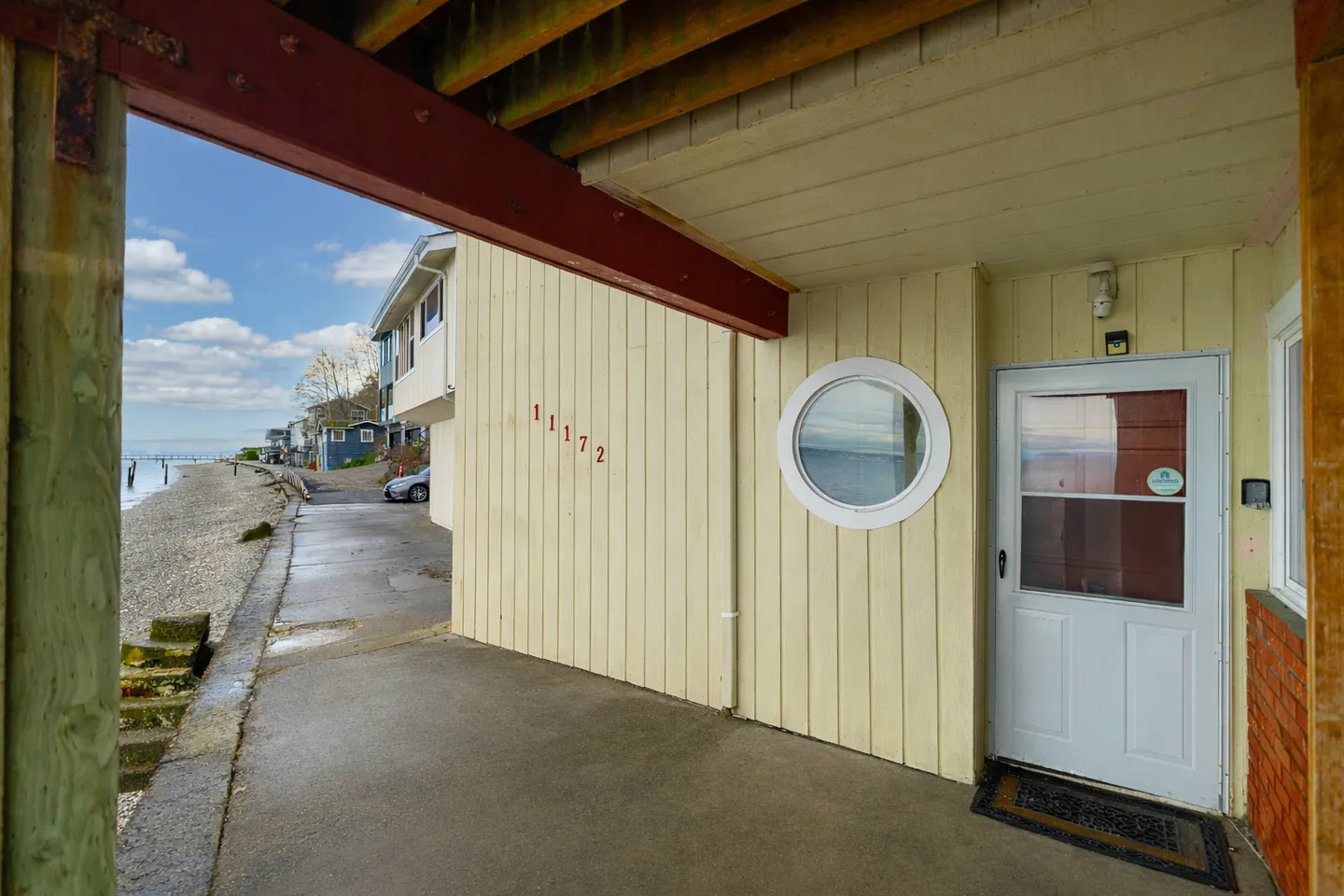 Covered concrete entryway of a light yellow home with a round window next to a shoreline street.