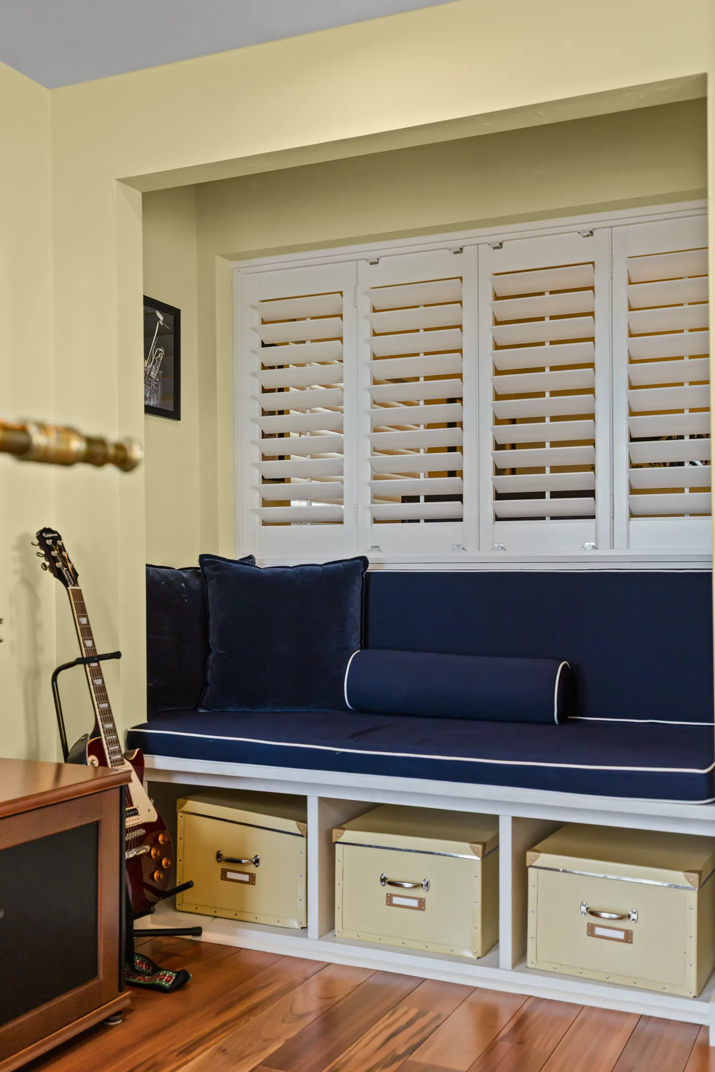 A built-in reading nook bench with dark blue cushions, pillows, white window shutters, and storage cubbies beneath holding cream boxes.