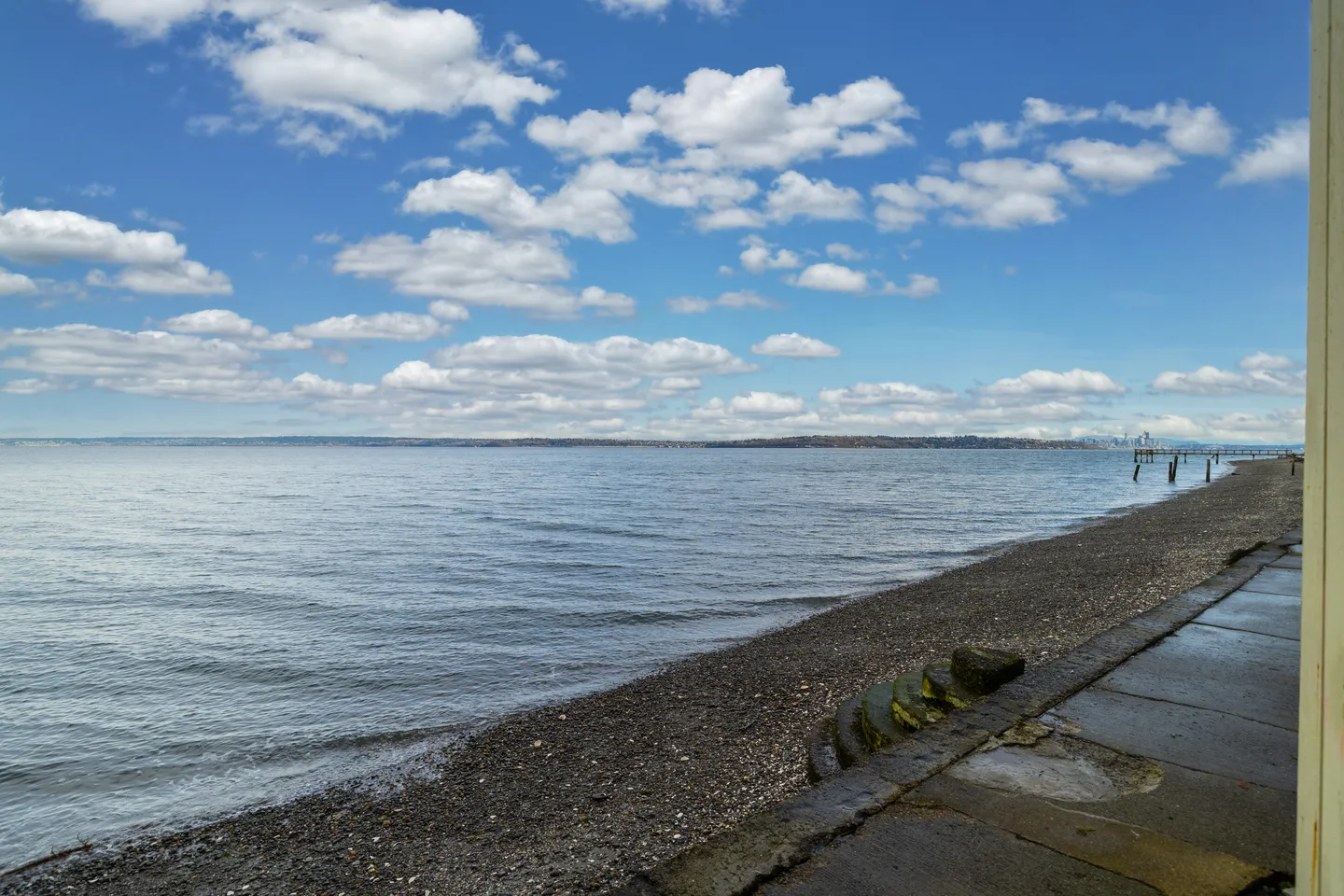 A wide view of a pebble shoreline and calm open water under a bright blue sky filled with white clouds.