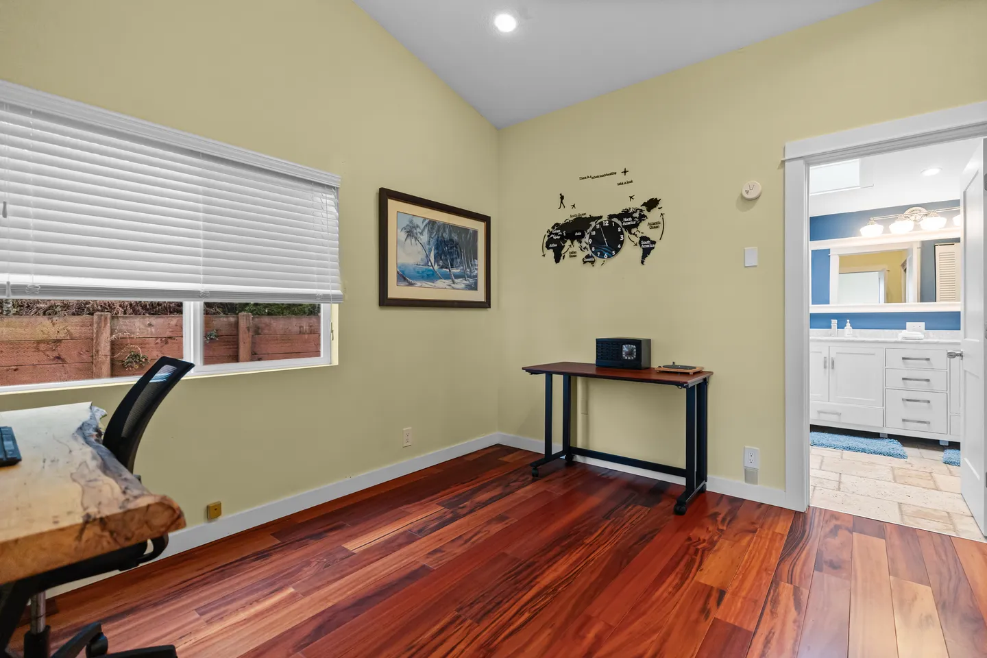 Workspace area with rich wood floors, a natural-edge desk, small side table, and a doorway leading into a white bathroom.