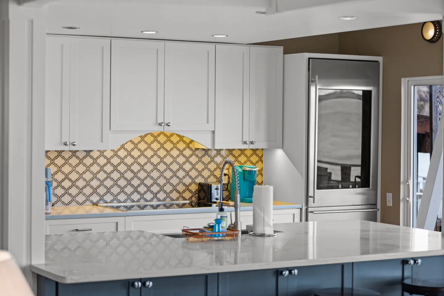 Kitchen counter and island with white cabinets, patterned tile backsplash, and stainless-steel refrigerator.