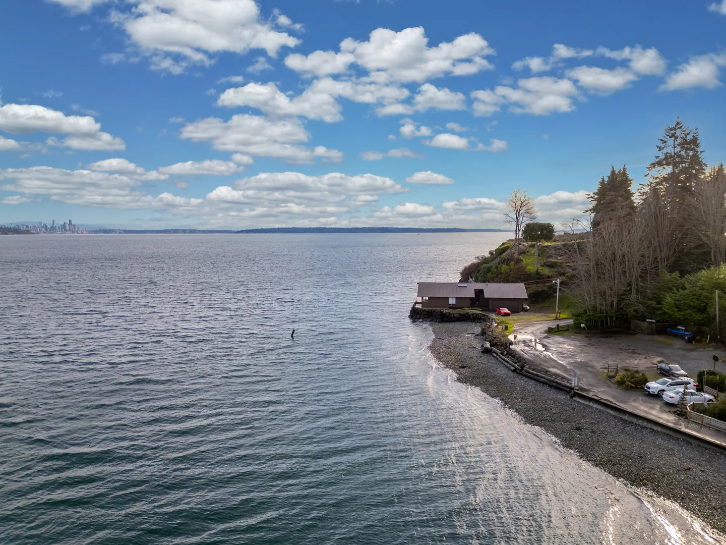Aerial view of a coastal house situated on a rocky shoreline with expansive water views under a partly cloudy sky.