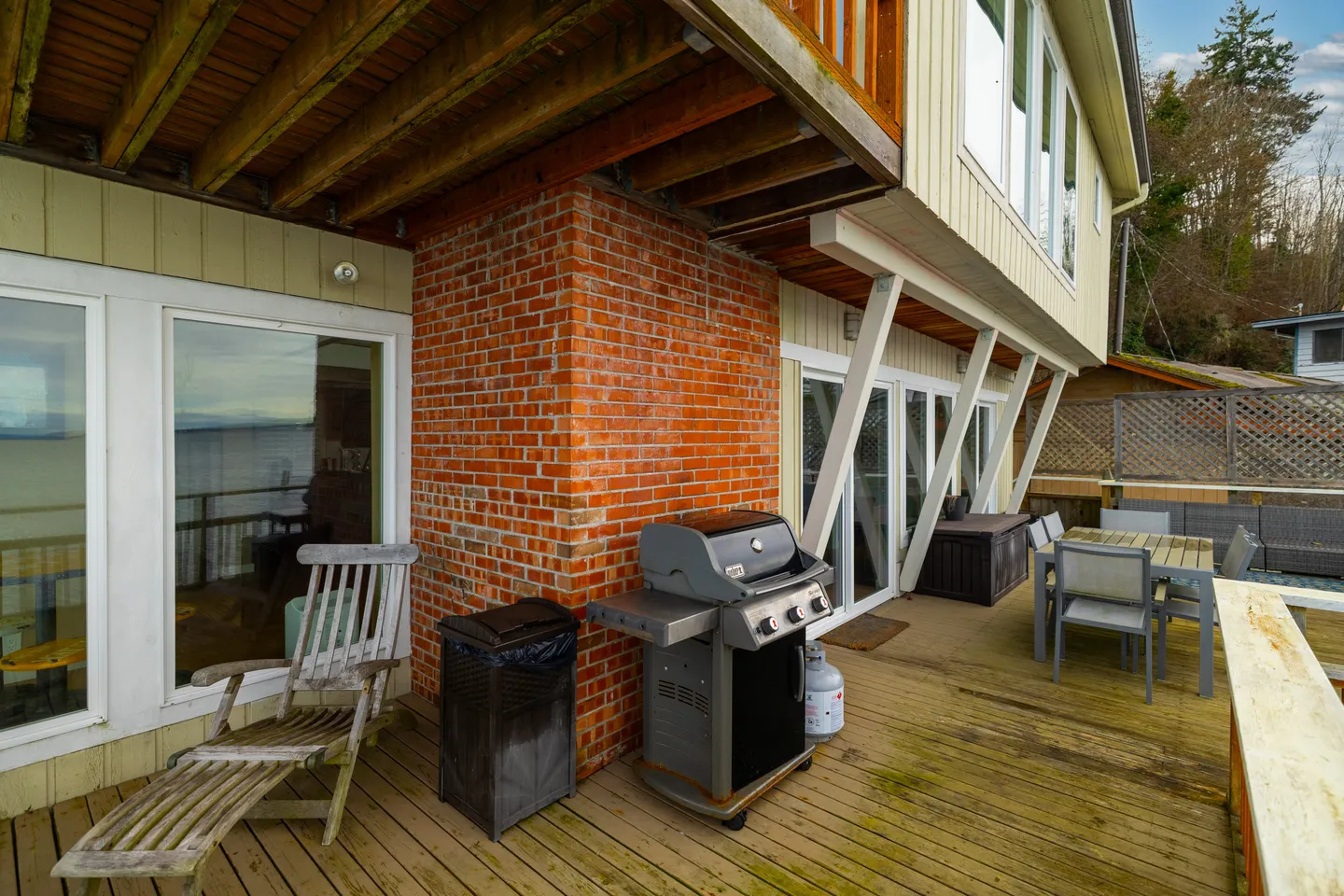 Lower wooden deck of a home featuring a gas grill, lounge chair, brick column, and an outdoor dining set under an upper-level overhang.