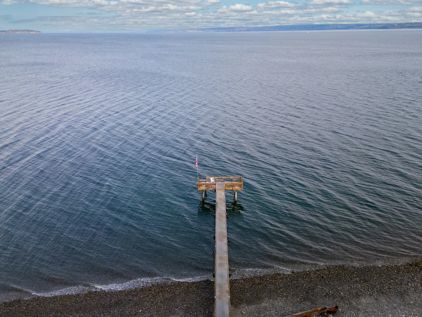 Aerial view of a long pier extending from a pebbled shoreline into calm open water under a partly cloudy sky.