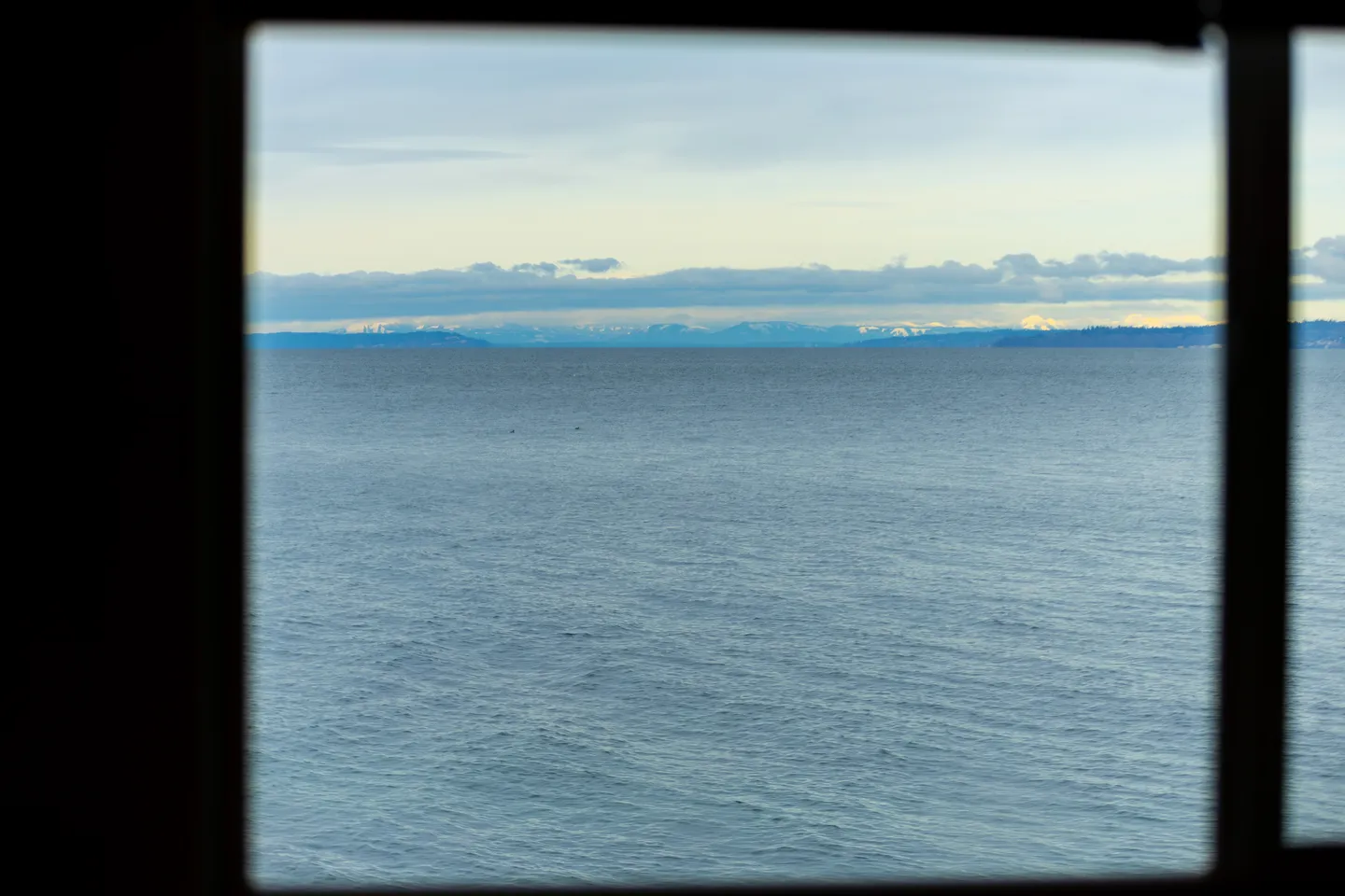 A wide view of the ocean and distant snow-dusted mountains seen through a window frame.