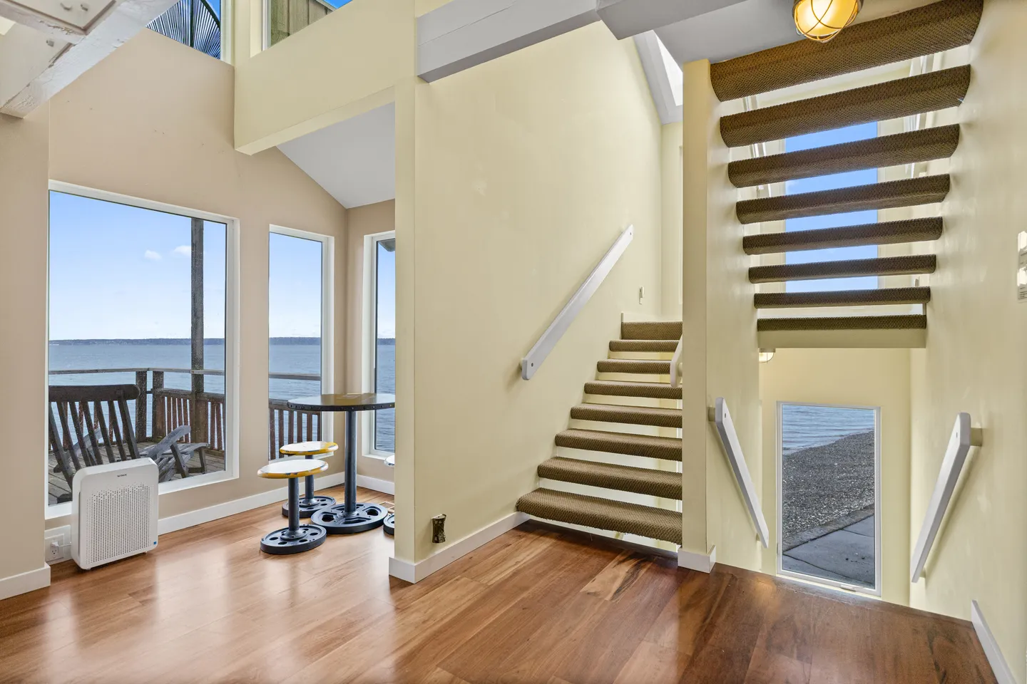 Open indoor entryway and staircase with light yellow walls, wood flooring, and large windows looking out onto water and an outdoor deck.