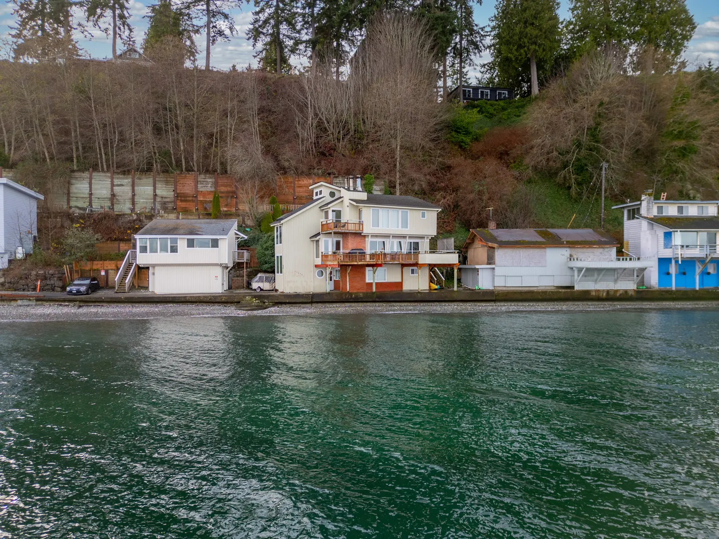 Elevated view from the water showing a multi-story waterfront home with multi-level decks along a shoreline beneath a wooded hillside.