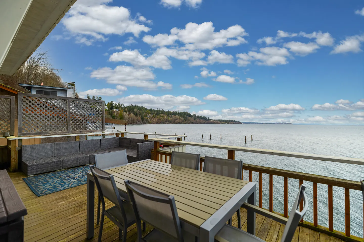Outdoor wooden deck overlooking a large body of water under a partly cloudy blue sky, equipped with a dining table, chairs, and a sectional couch.
