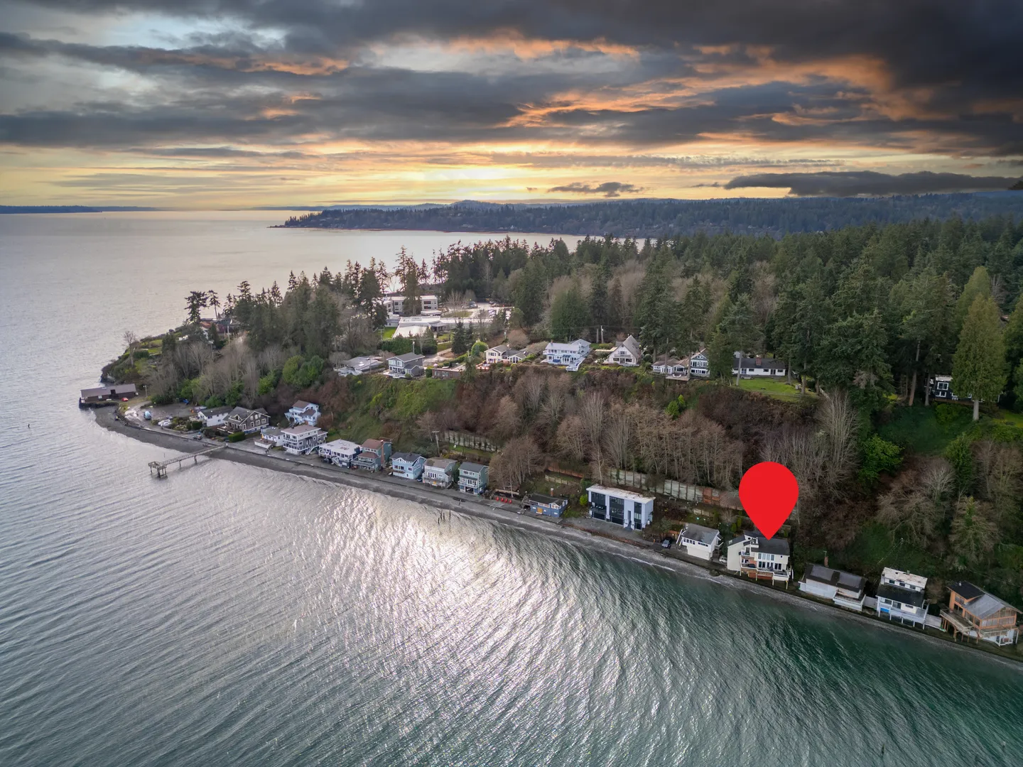 Aerial view of a coastal shoreline with beachfront homes, forested bluffs, and a red location pin marking one property at sunset.