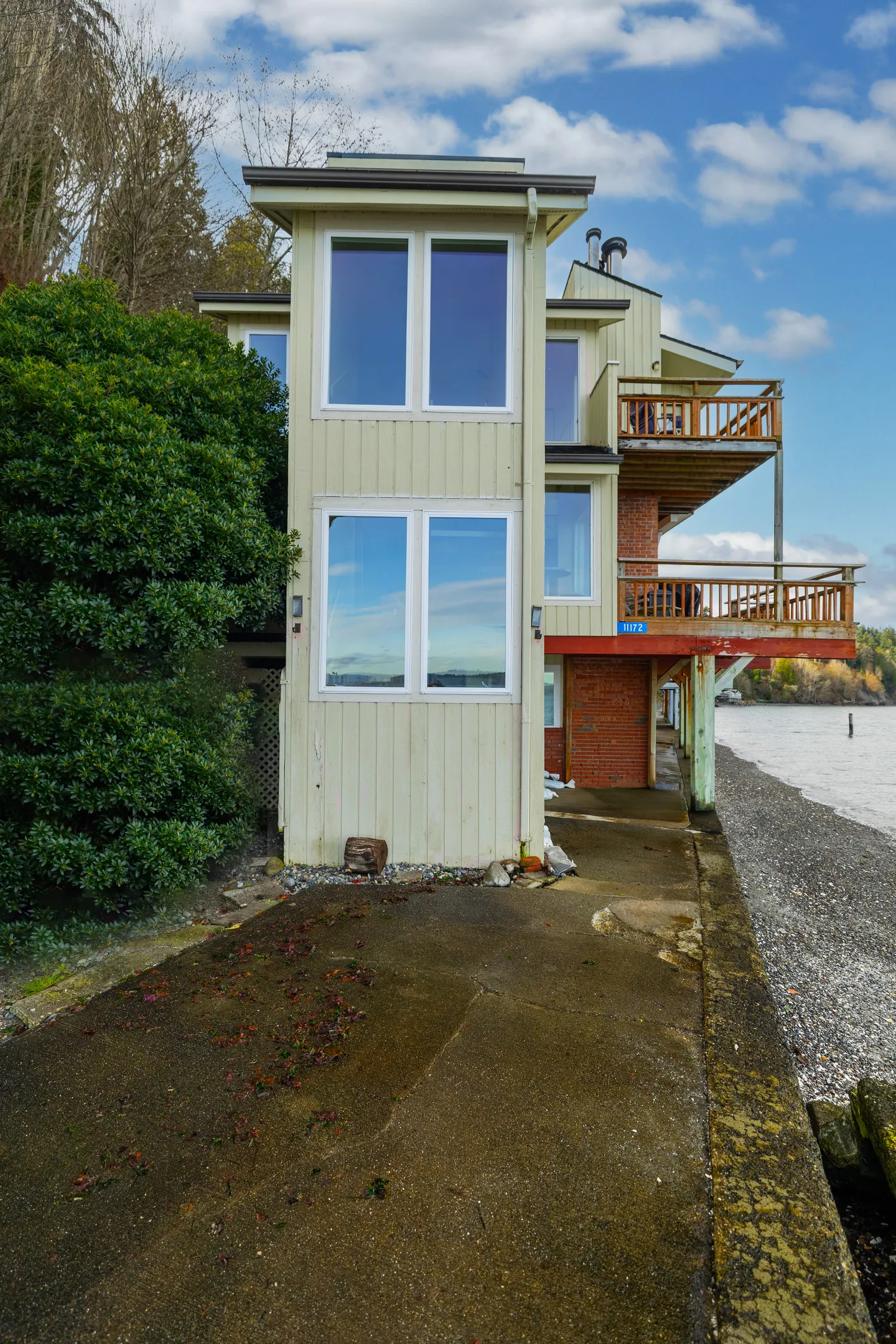 Exterior of a multi-story waterfront home with light siding, wooden balconies, and a concrete patio next to a rocky beach.