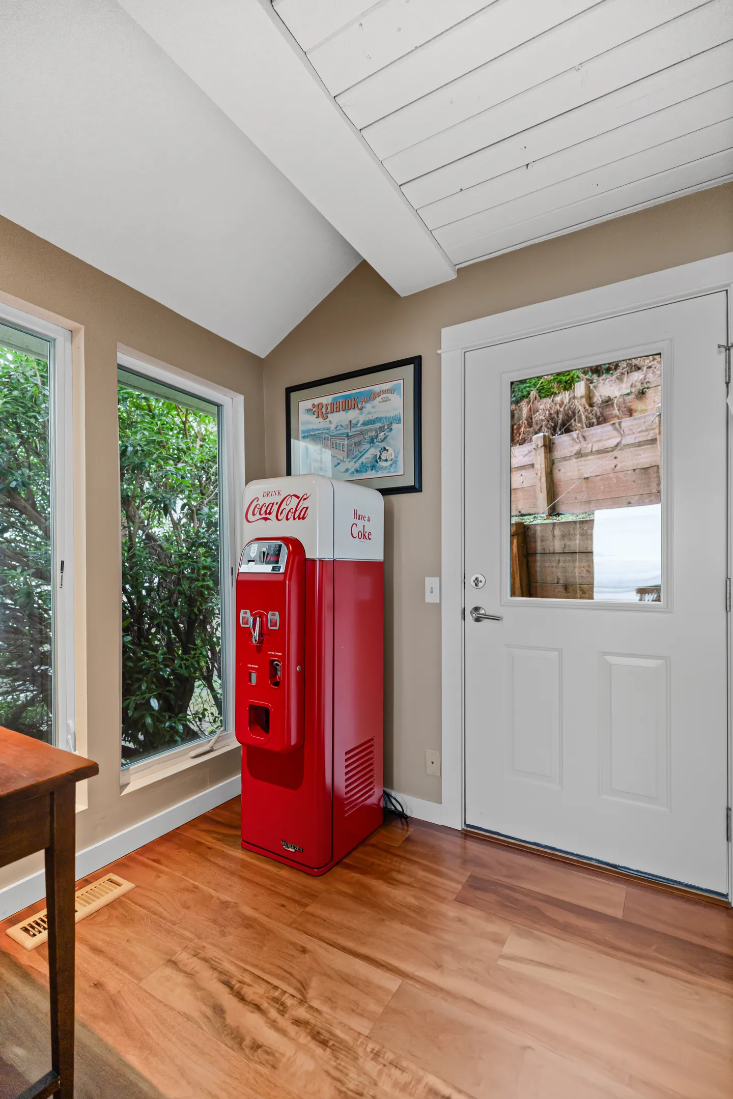A corner of a room featuring a vintage-style red and white Coca-Cola vending machine next to a white exterior door and windows.
