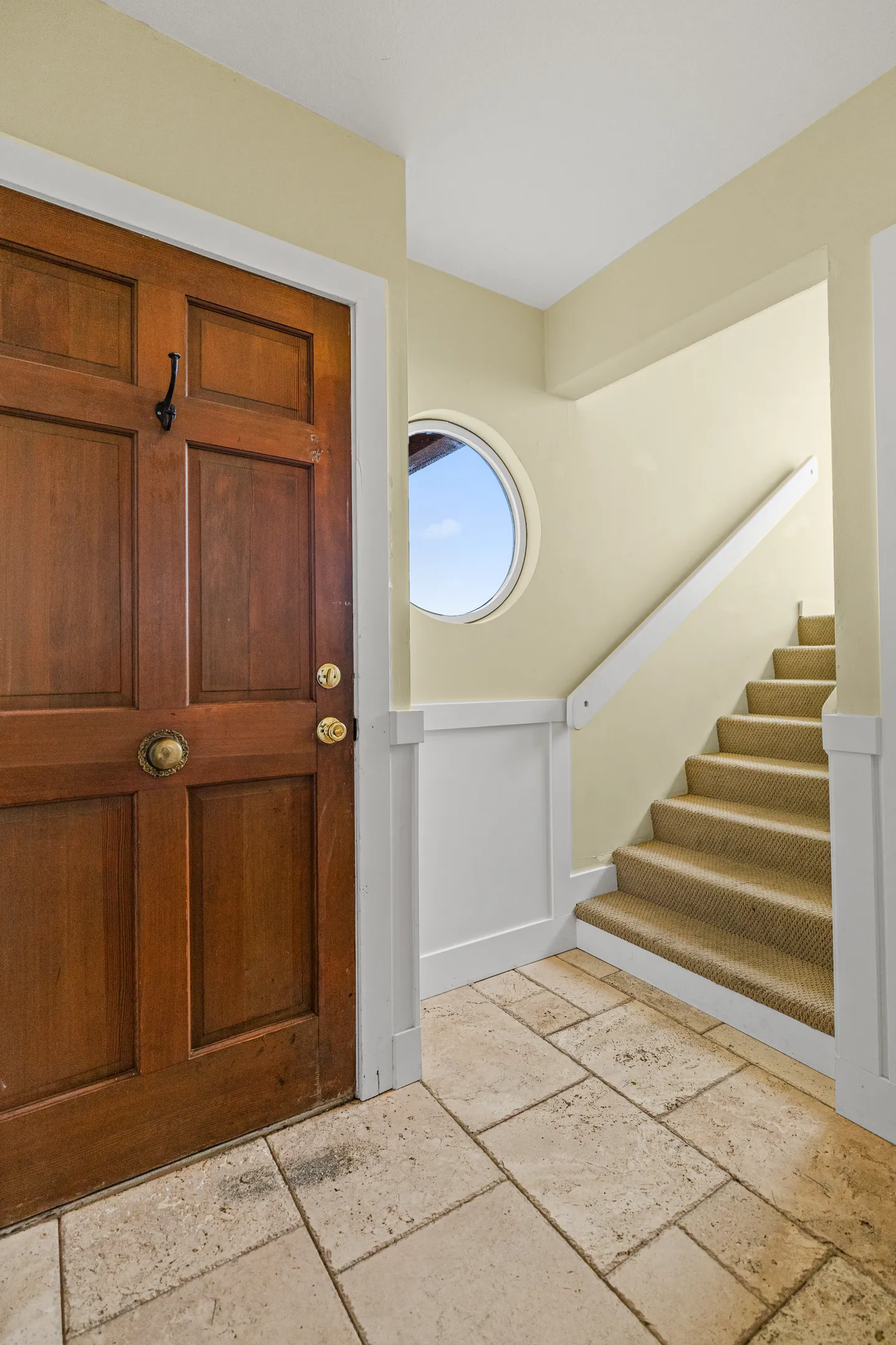 Entryway featuring a solid wood front door, tiled flooring, a small round window, and carpeted stairs leading up.