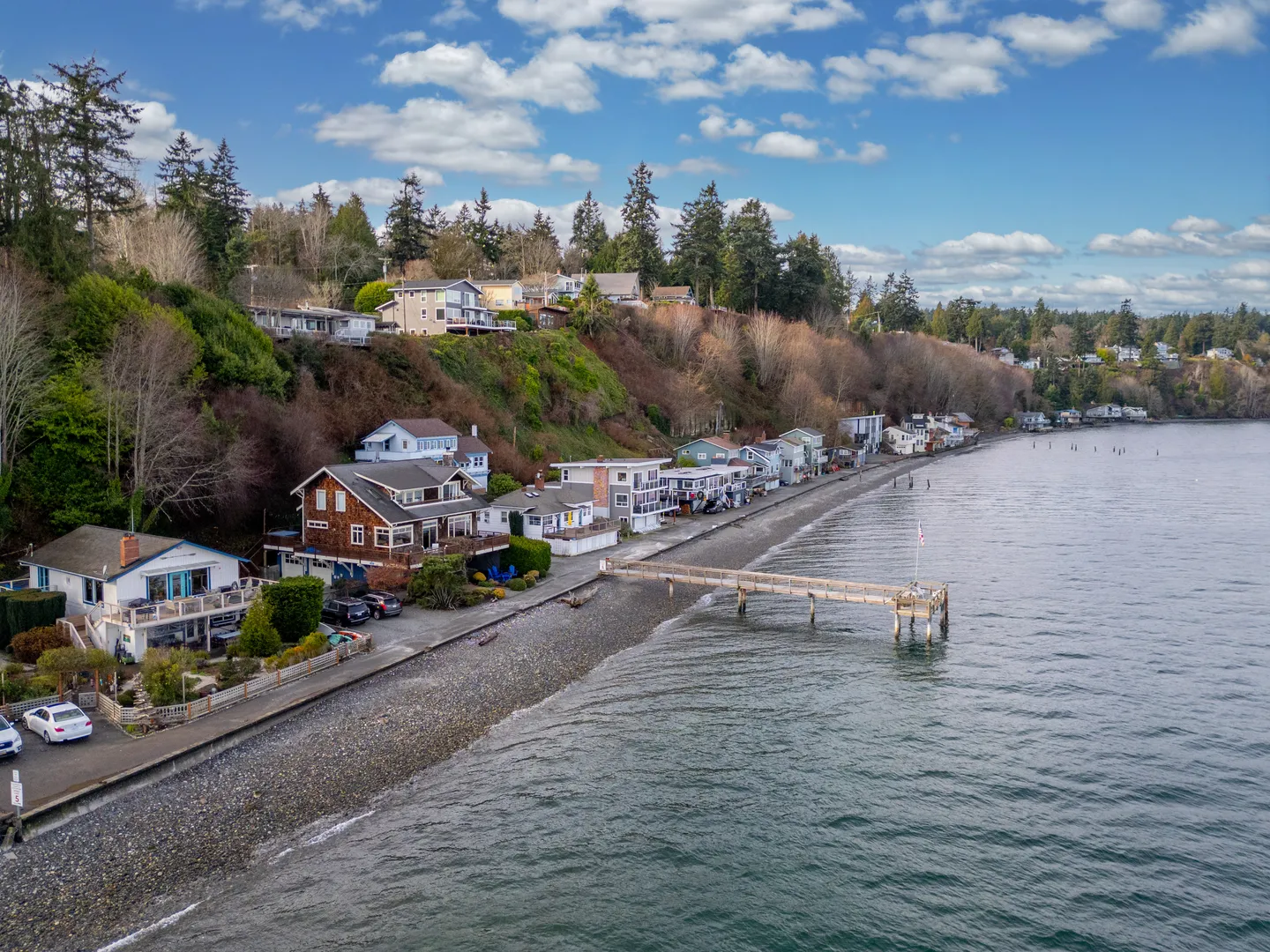 Aerial view of waterfront houses along a gravel shoreline with a wooden pier extending into the water.