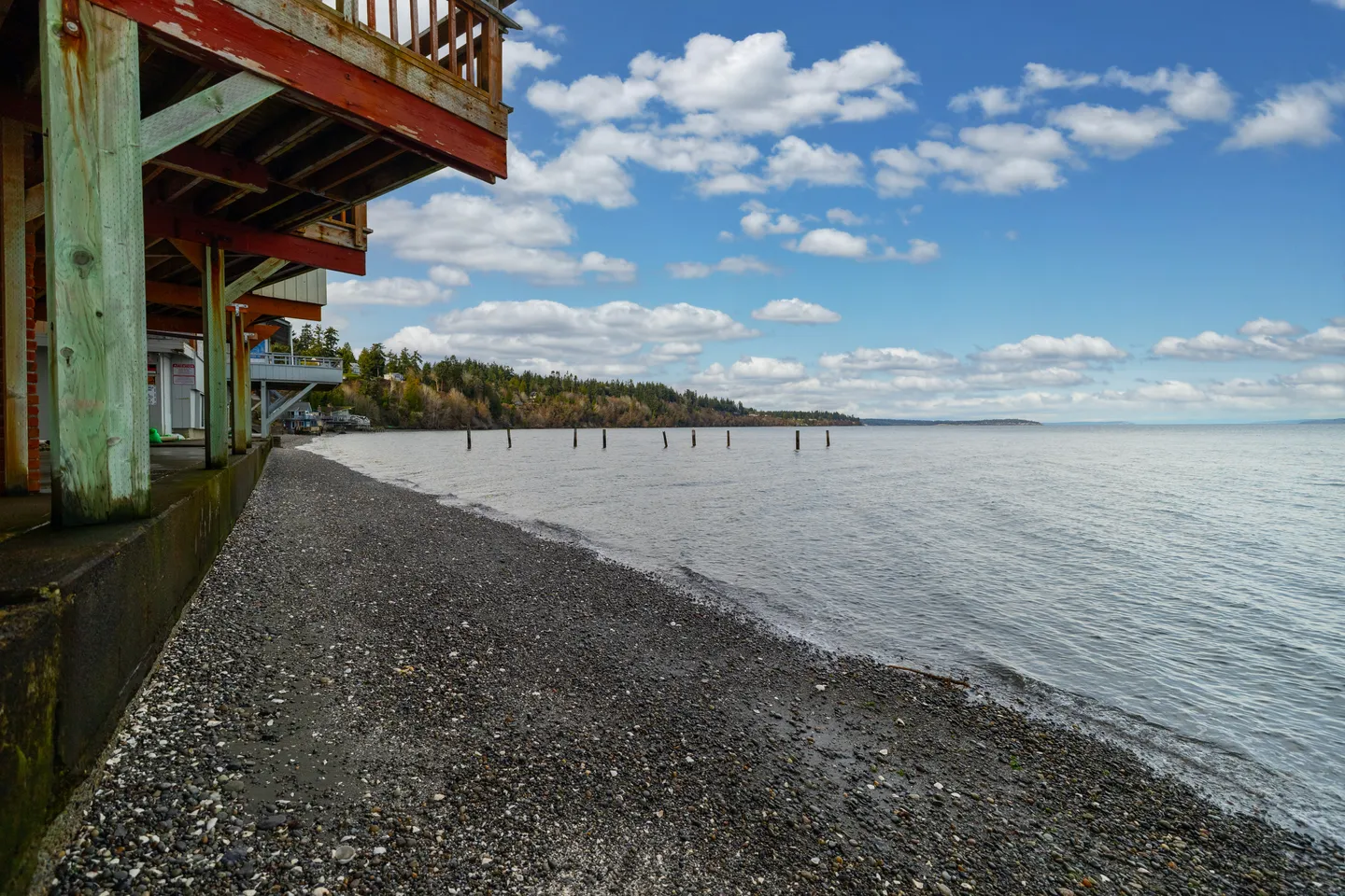 View from beneath an elevated deck looking out along a pebble beach and calm water under a partly cloudy blue sky.