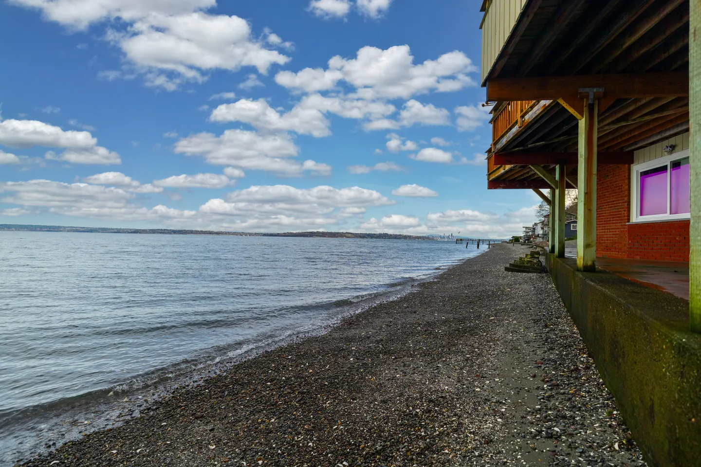 A gravel and shell beach beside a calm body of water next to the lower level of a waterfront building on a partly cloudy day.