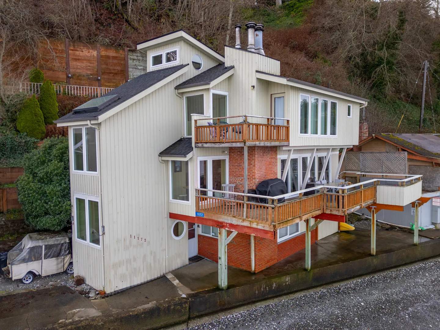 Exterior view of a multi-story light-colored house built against a hillside with multiple wooden decks and a seawall.