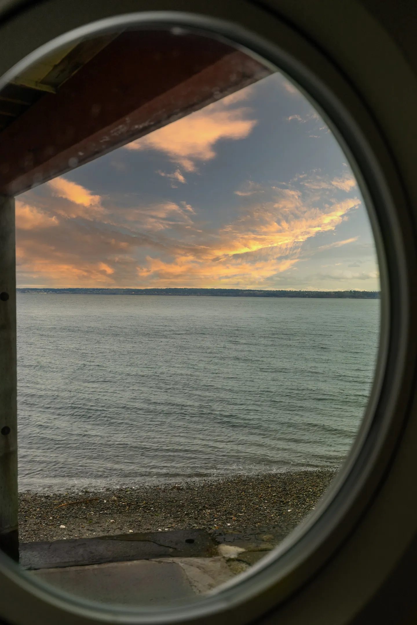 A view of the ocean shoreline and sunset sky framed through a round porthole window.