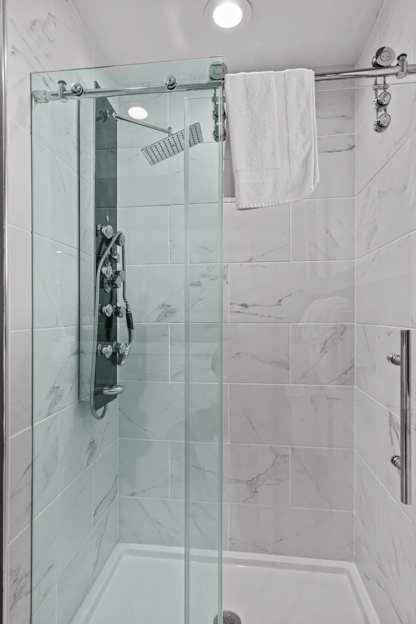 A walk-in shower with sliding glass doors, white marble-patterned tile, and a multi-function shower tower panel.