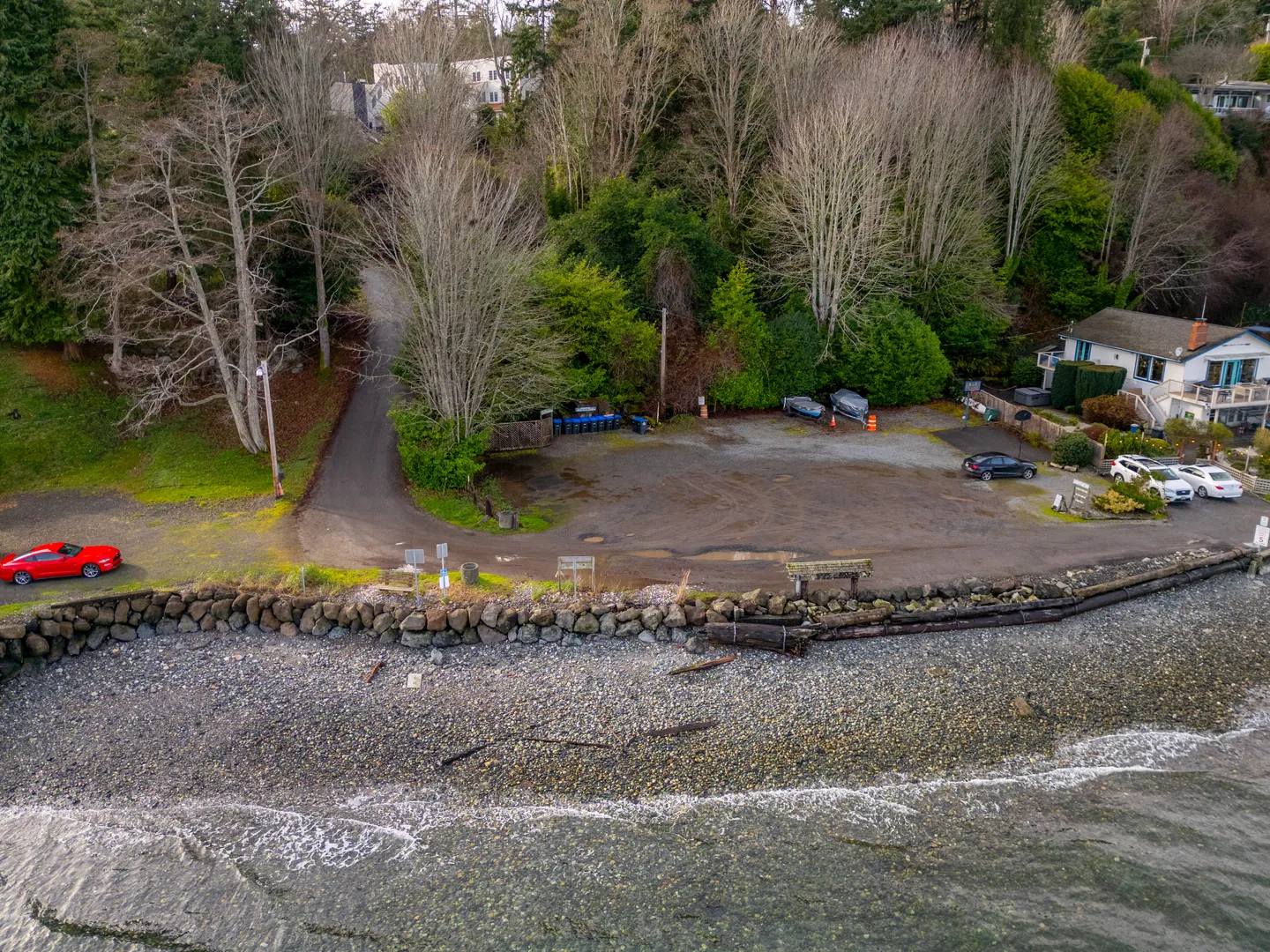 Aerial view of a coastal parking area, rocky beach shoreline, and access road bordered by trees.