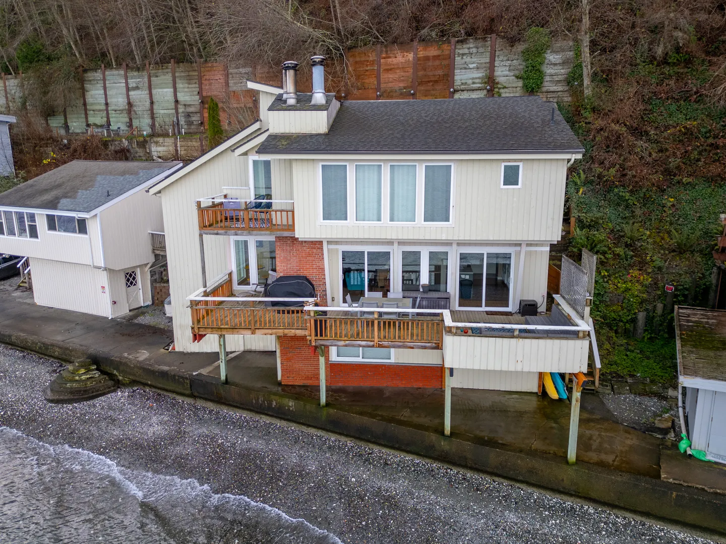 Elevated view of a waterfront two-story home with light-colored siding, multiple wooden decks, and large windows directly overlooking a shoreline.