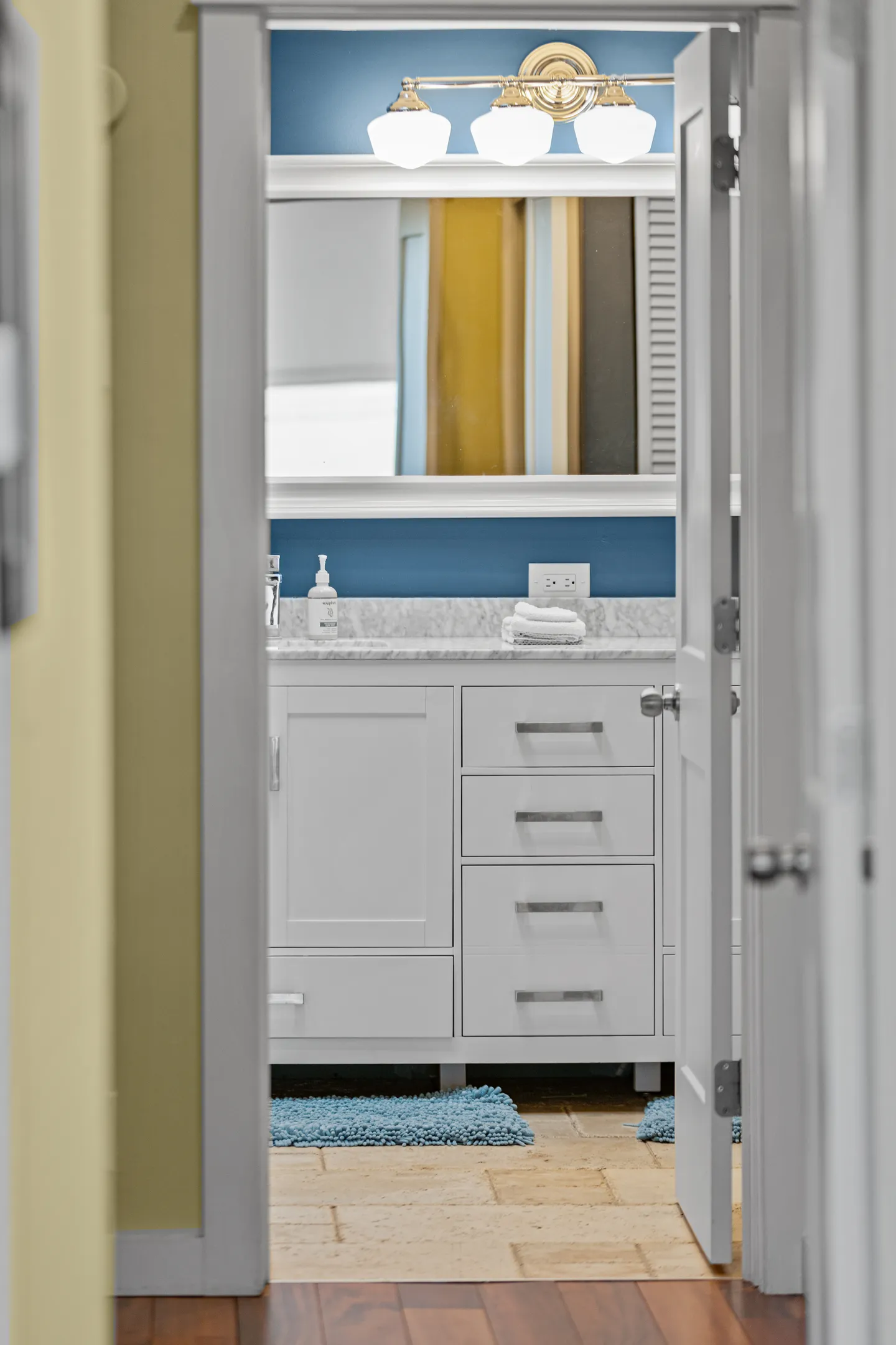 View through an open doorway of a bathroom vanity with white cabinets, blue wall, and vanity mirror.