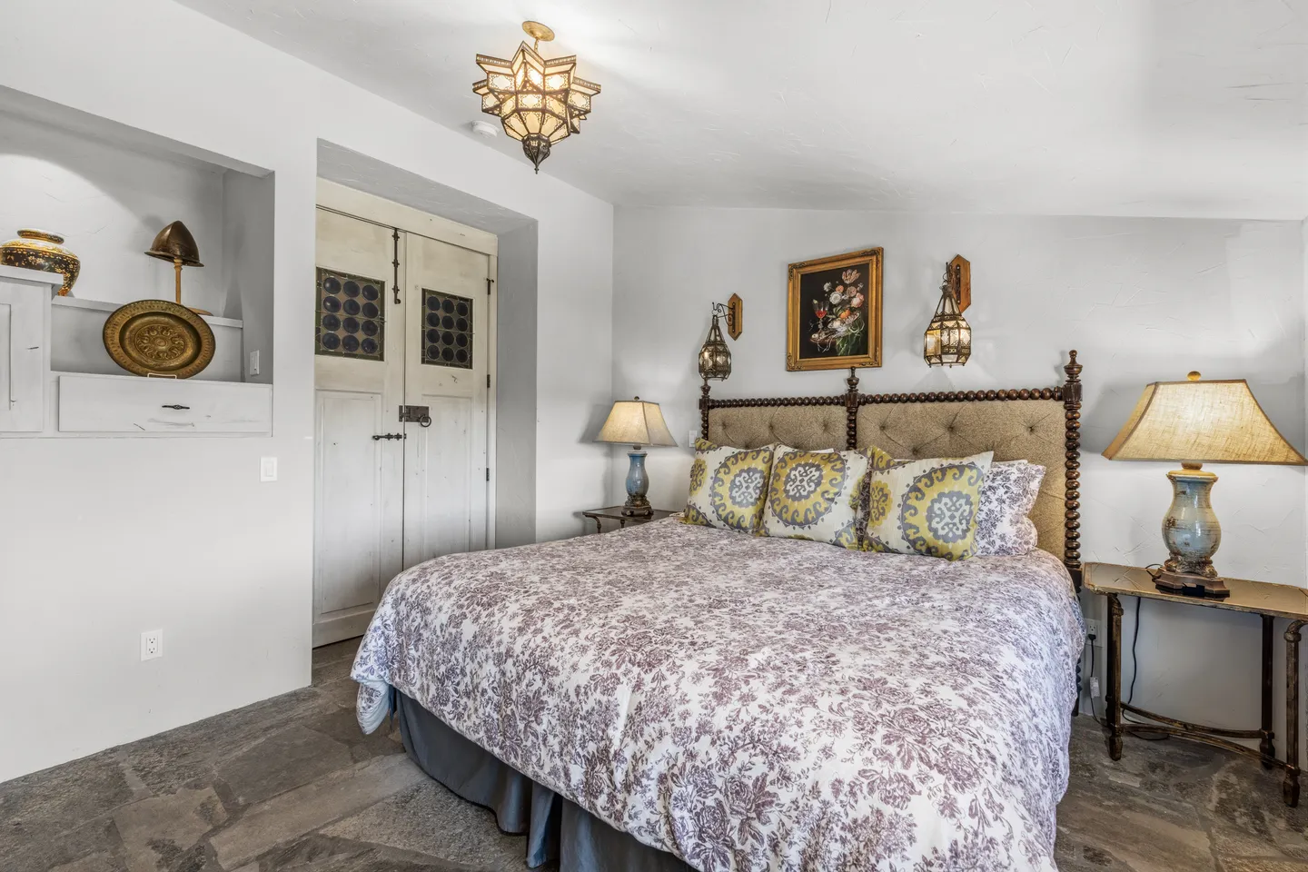 Bedroom with a patterned bedspread, tufted headboard, stone tile floor, and rustic accent lighting.