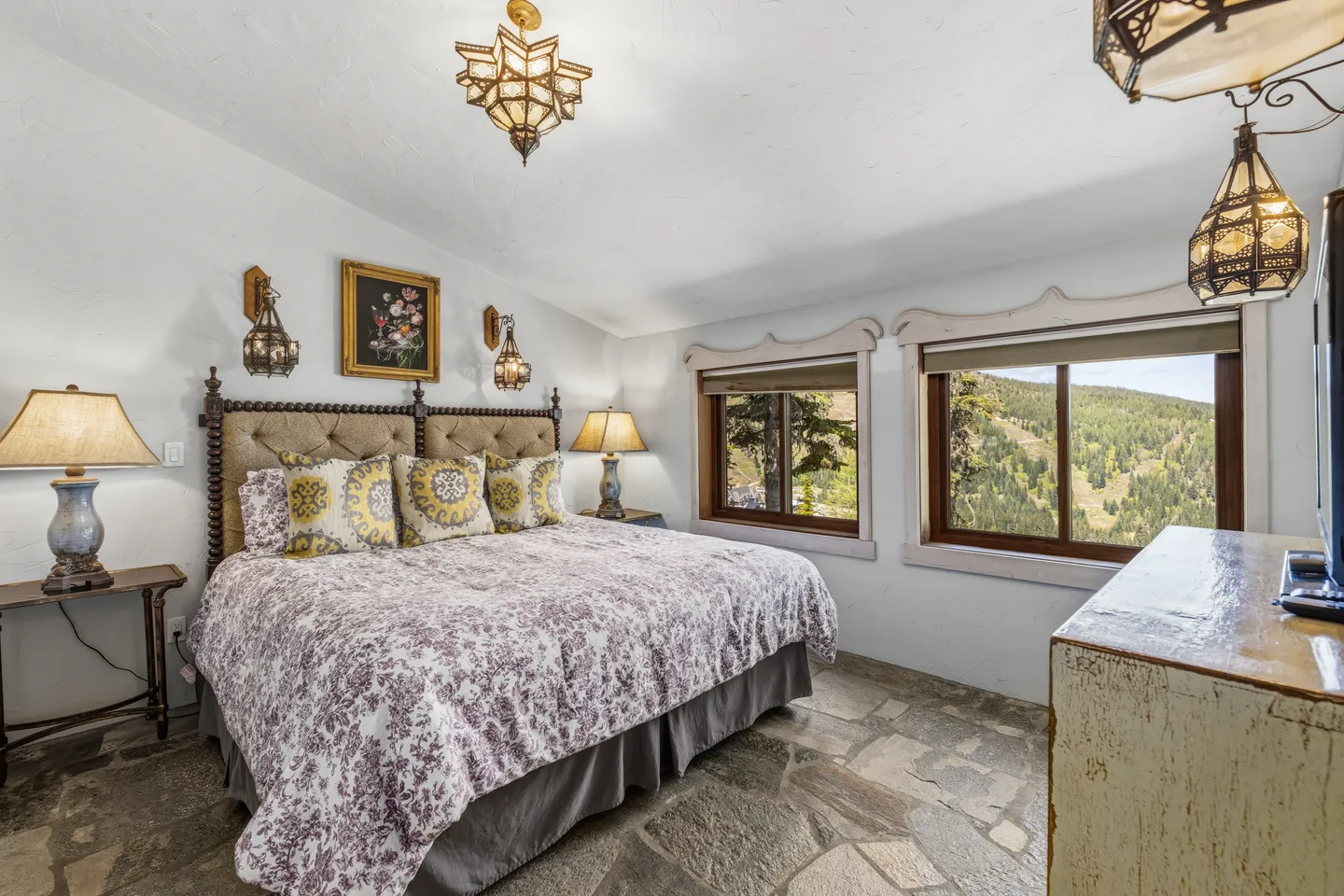 Bedroom with stone tile flooring, an upholstered bed, decorative hanging lanterns, and windows looking out to mountains.