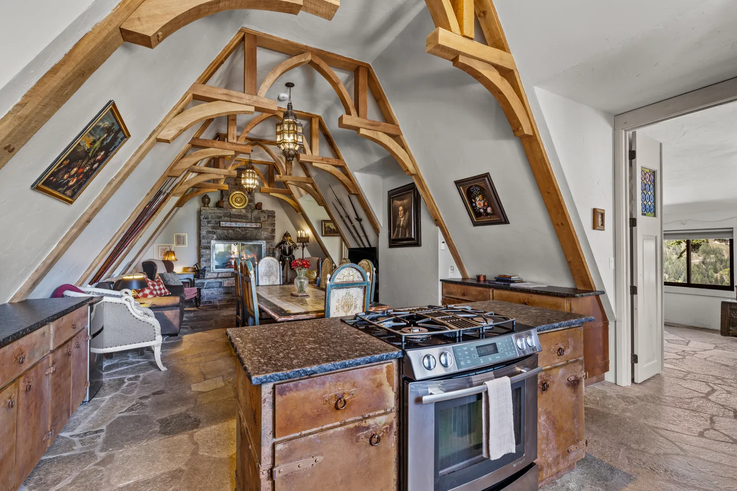 Open-concept interior featuring stone tile floors, prominent curved wood ceiling trusses, kitchen island with stove, dining table, and stone fireplace in the background.