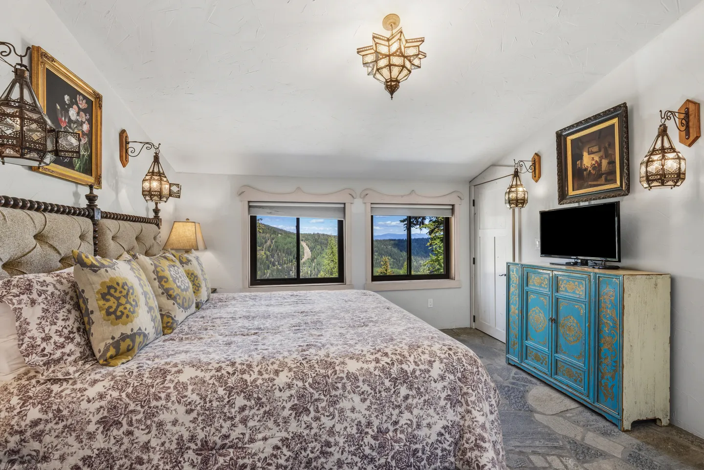 Bedroom featuring a made bed with an upholstered headboard, hanging lanterns, and windows looking out to mountains.