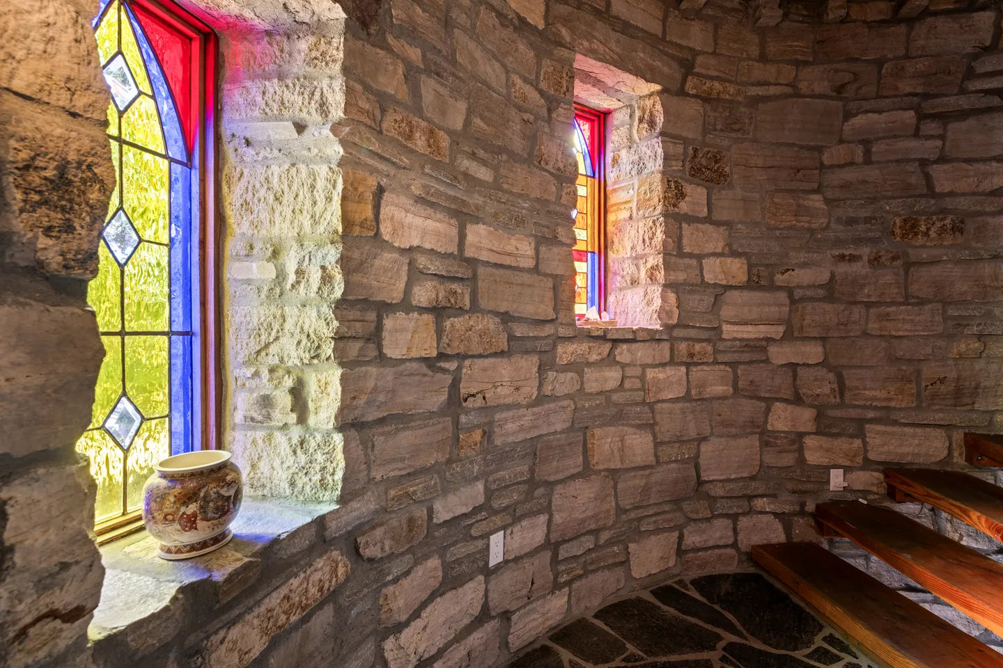 Curved stone wall along an interior staircase featuring colorful stained-glass windows and a decorative vase on the deep windowsill.