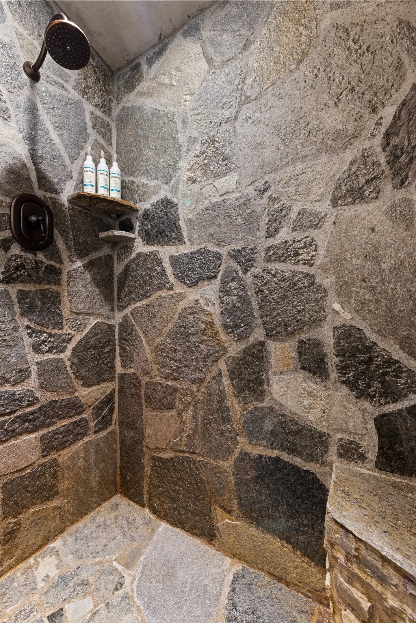 Walk-in shower stall with natural stone walls, a dark metal showerhead, and small built-in corner shelves holding toiletries.