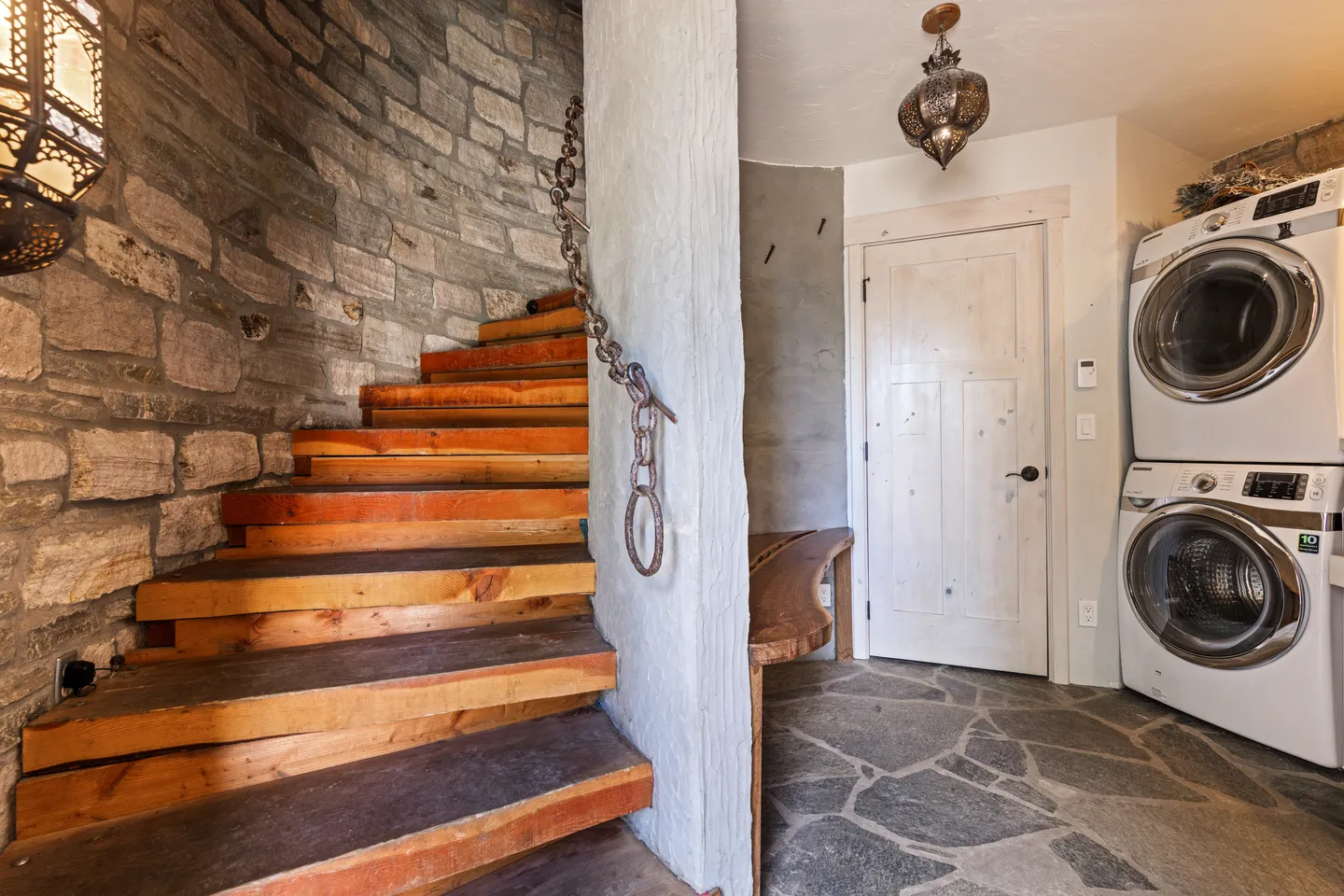A rustic interior hallway featuring stone flooring, a stacked washer and dryer, a wooden curved staircase with stone walls, and an ornate hanging lantern.