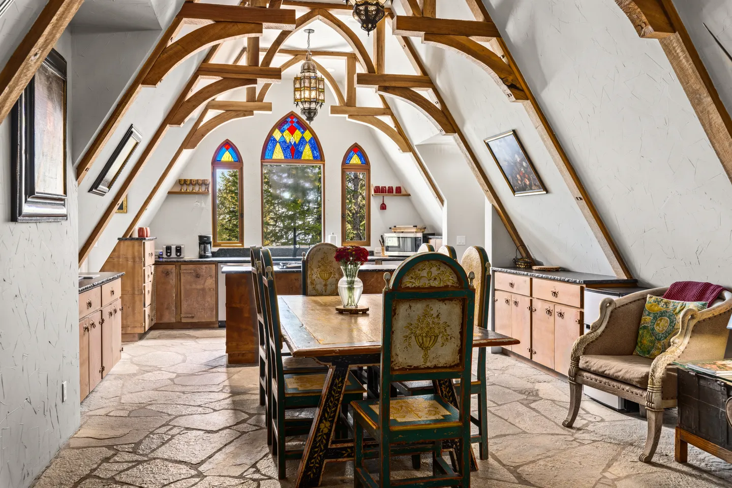 Dining area with vaulted timber beams, stone tile flooring, painted wooden chairs, and arched stained-glass windows leading into an open kitchen.