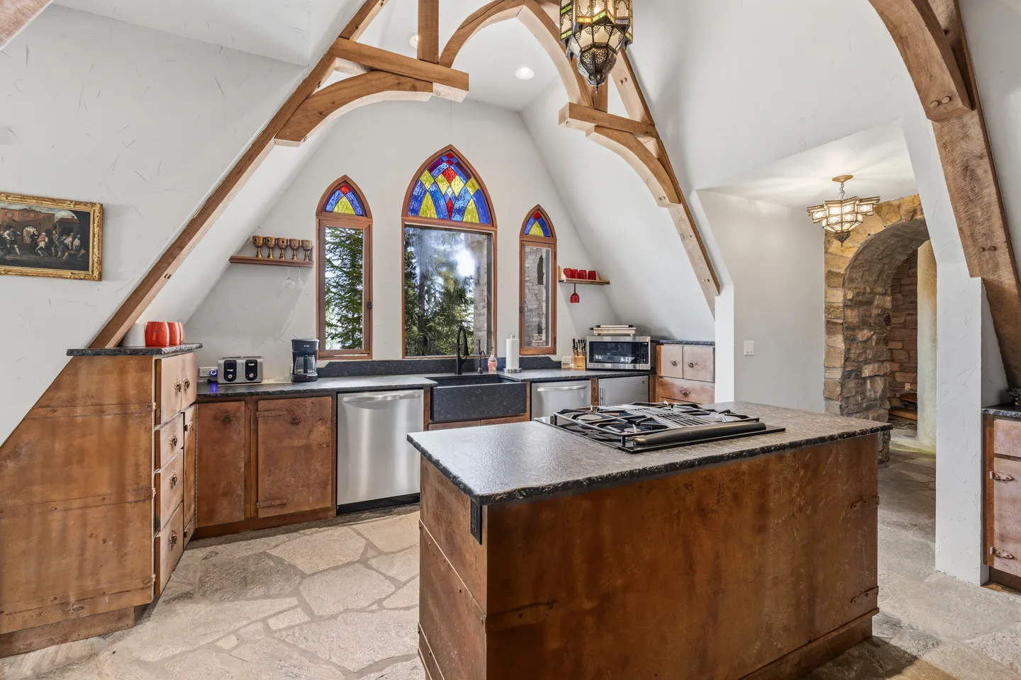 Kitchen with arched exposed beams, stained glass windows above the sink, stone flagstone flooring, and a center island with a cooktop.