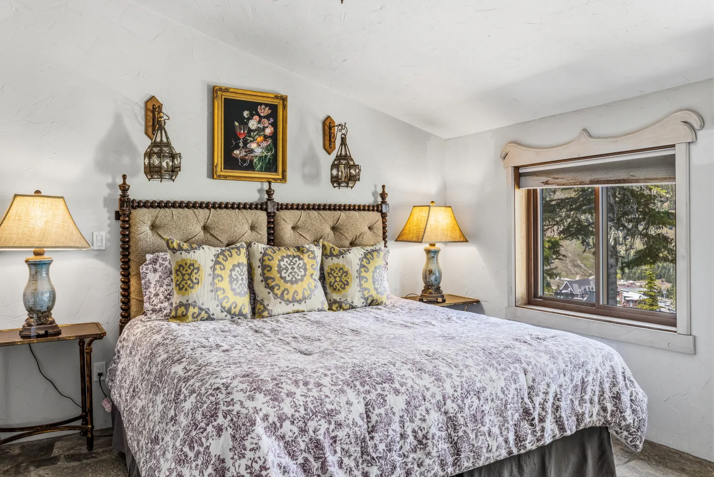 Bedroom with a patterned bedspread, tufted headboard, bedside lamps, and a window with mountain views.