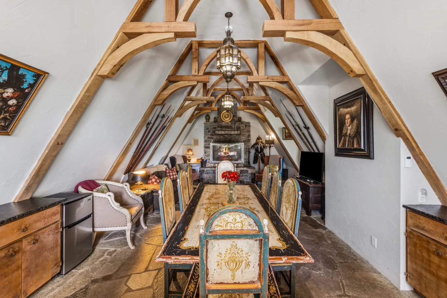 A-frame room with exposed wooden arched trusses, a long dining table, stone flooring, and a fireplace in the background.