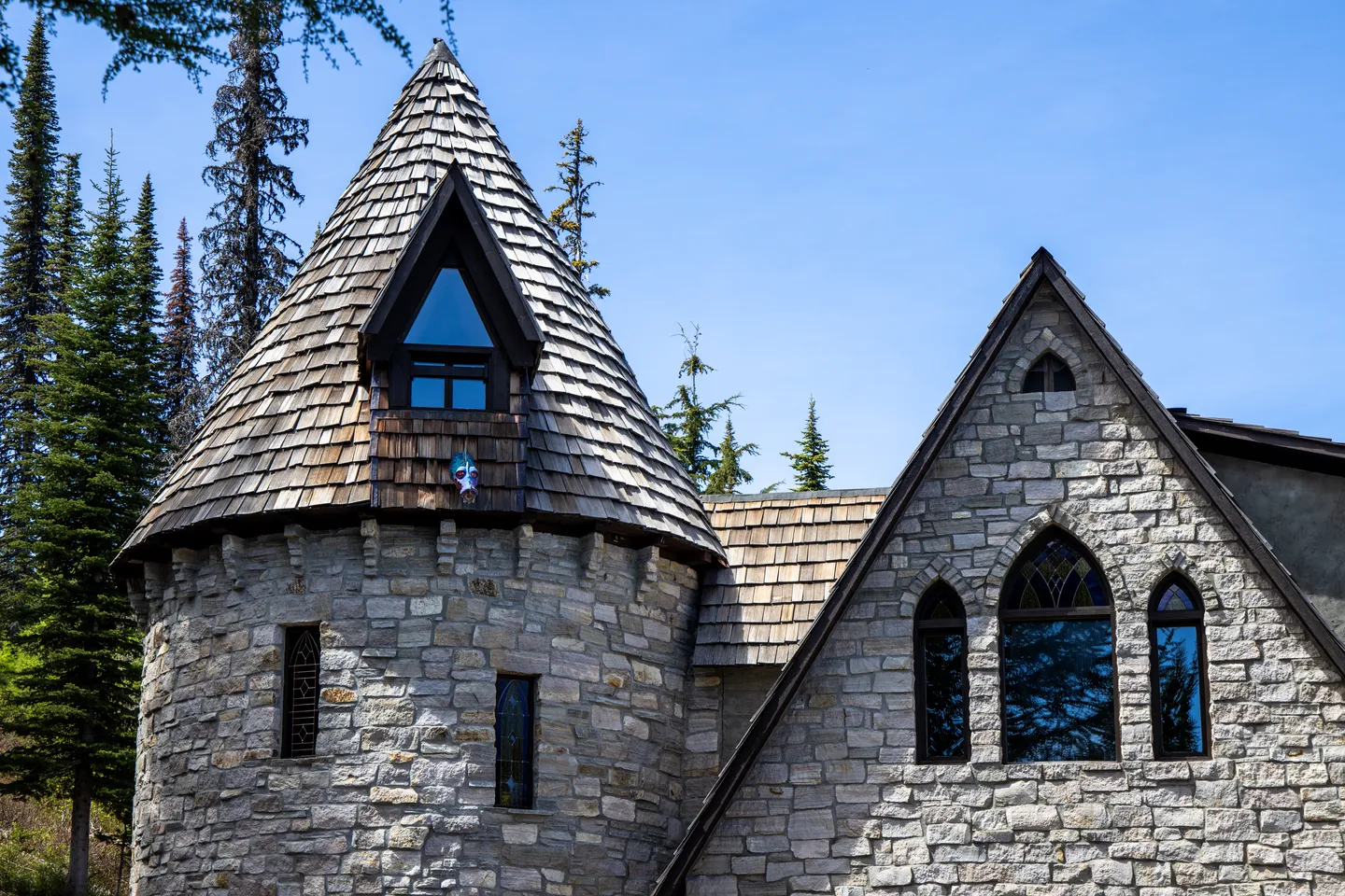 Exterior view of a stone turret with a wood-shingled conical roof alongside a stone gable wall under a clear blue sky.